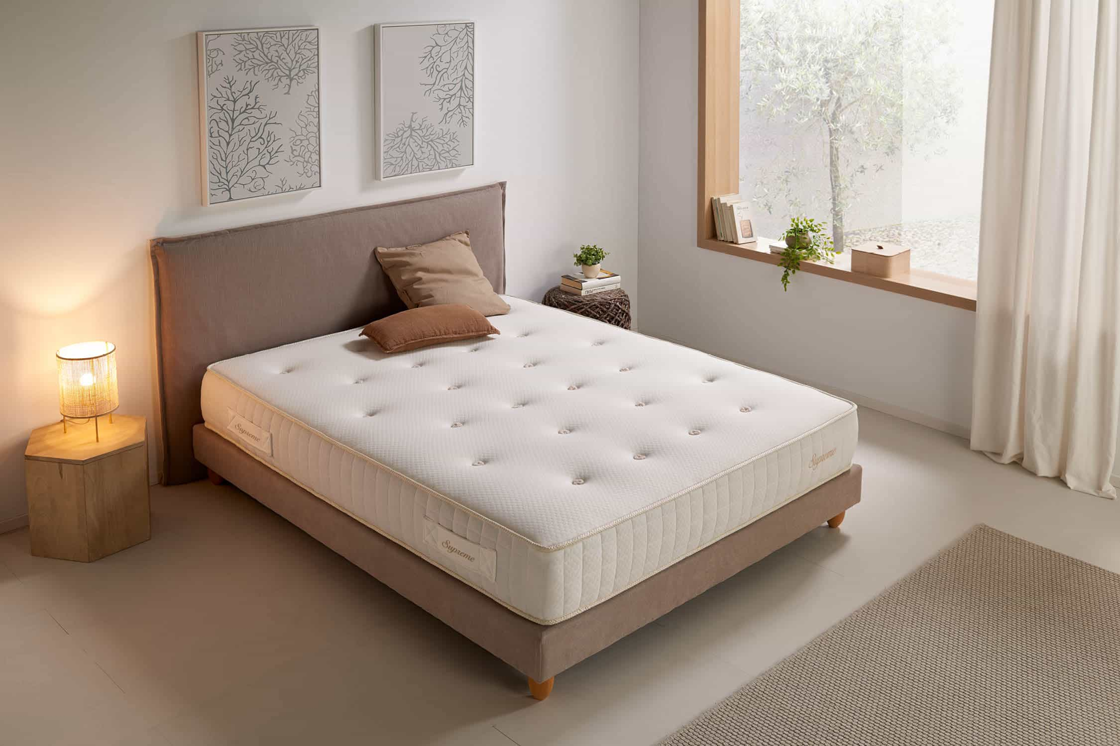 Cama con colchón y almohadas. Marco de cama beige. Colchón blanco con etiqueta Supreme. Ambiente de habitación.