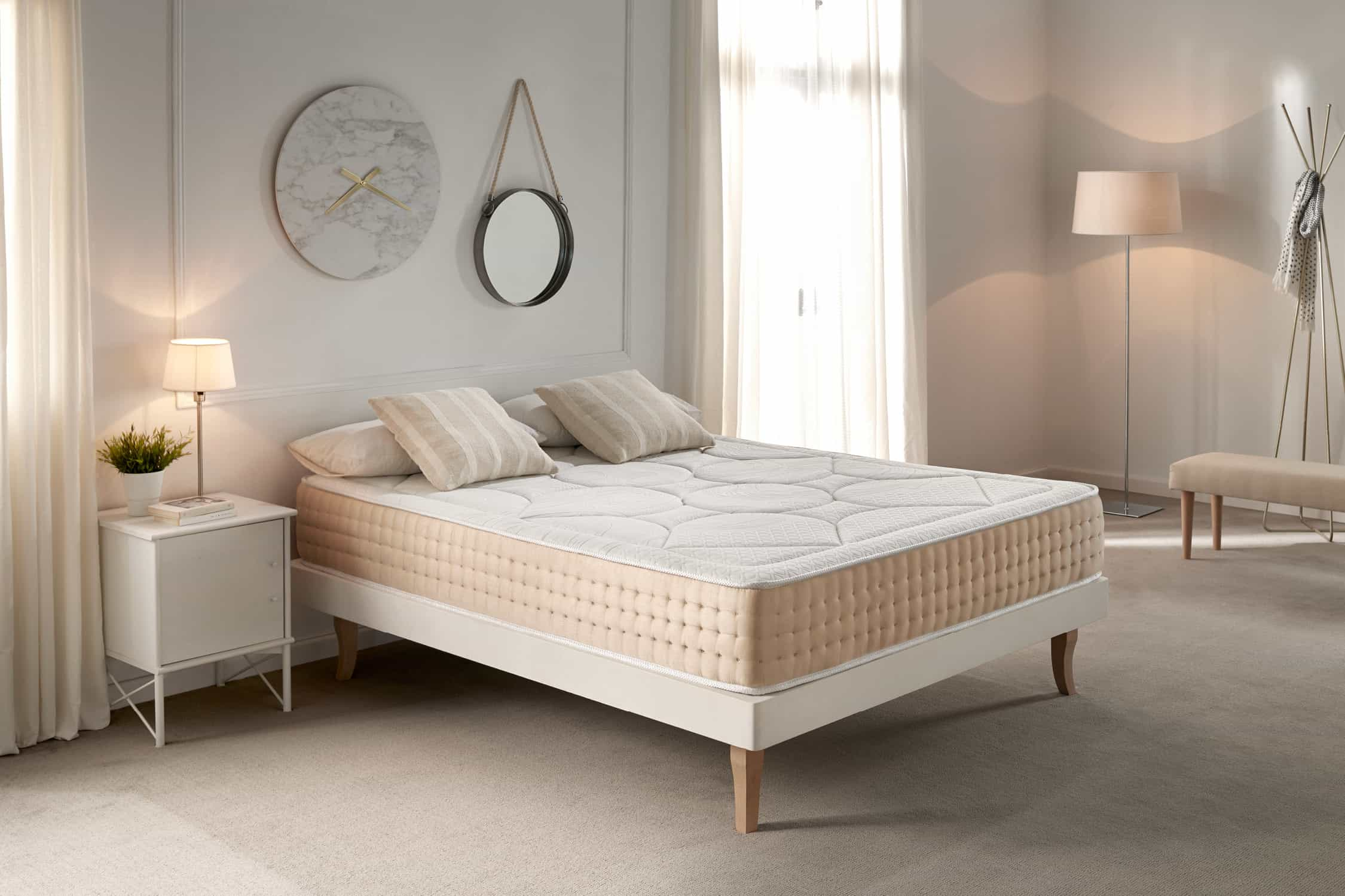 Interior de dormitorio con colchón en cama, mesita de noche, lámpara, almohadas. Colores y tonos neutros.