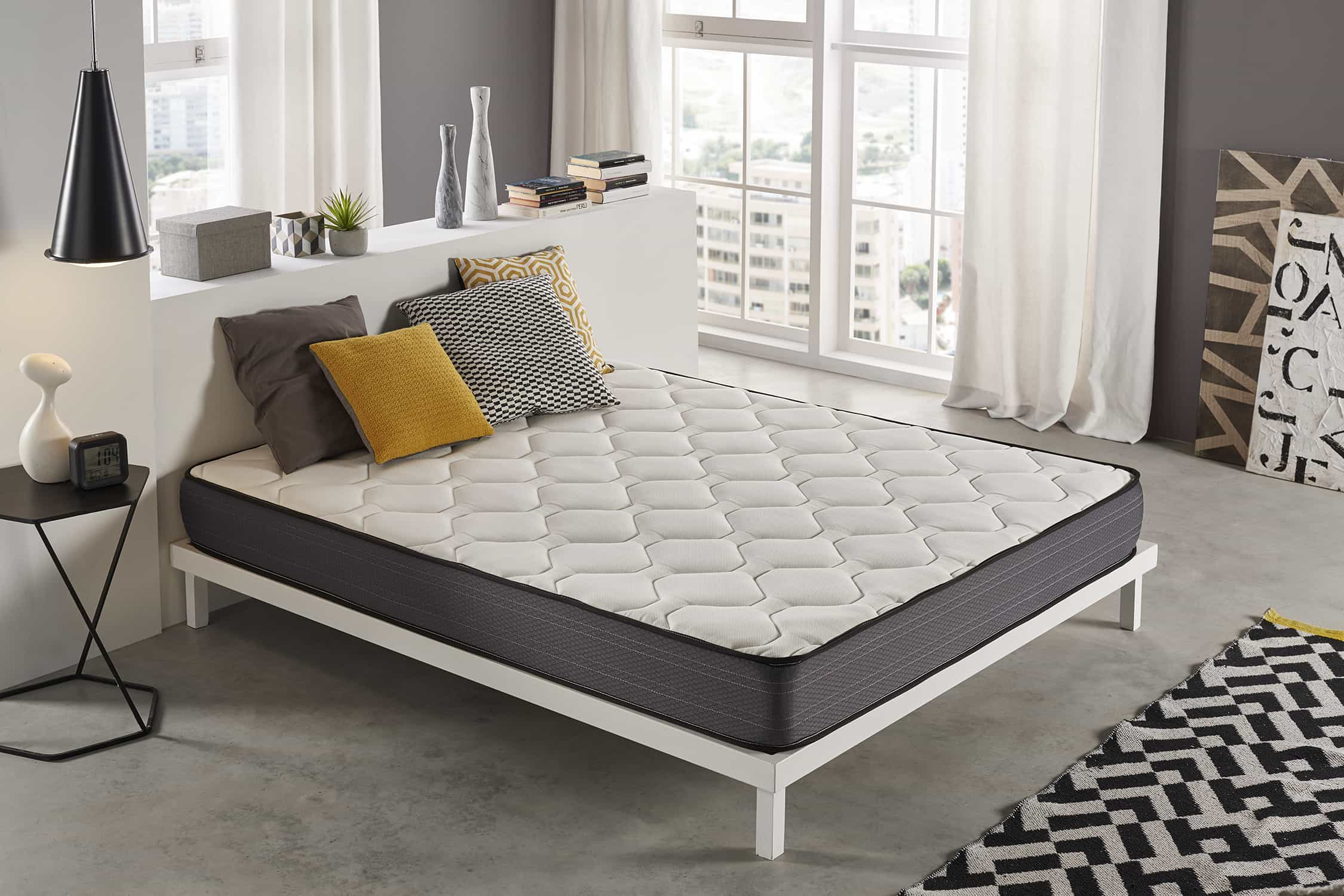 Colchón blanco con almohadas. Lámpara negra, mesa pequeña. Alfombra y pared blanca al fondo.