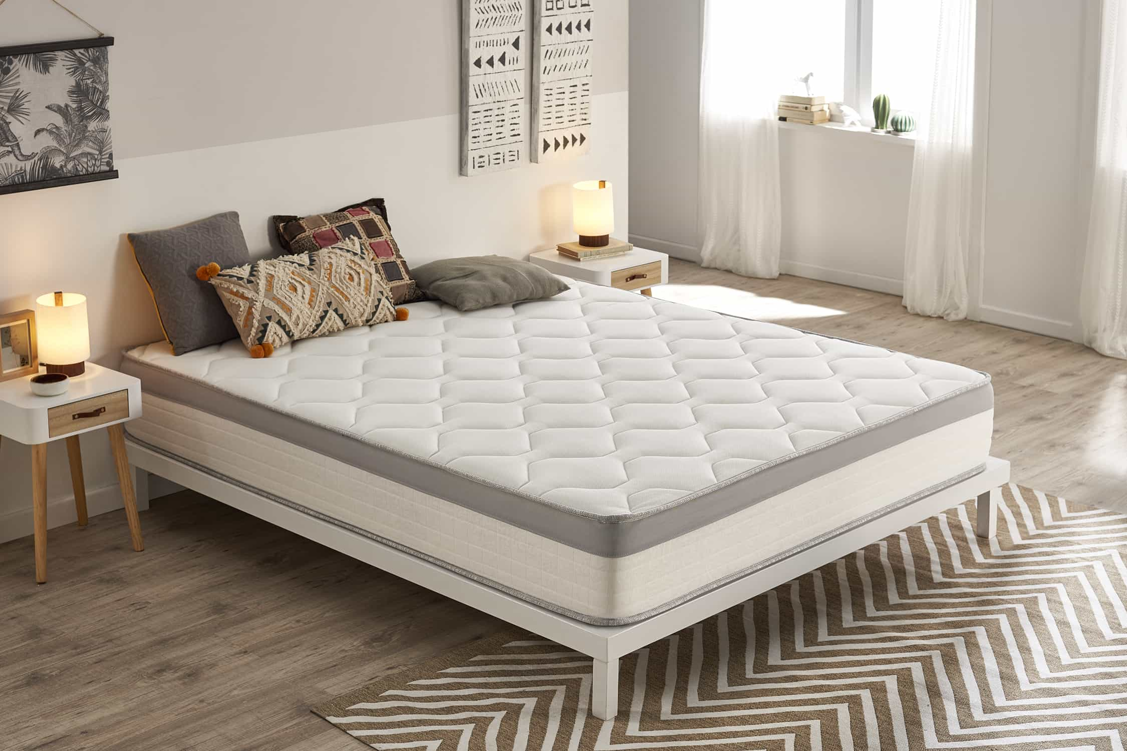 Colchón blanco con borde gris. Almohadas en la cama, mesitas de noche y una alfombra.