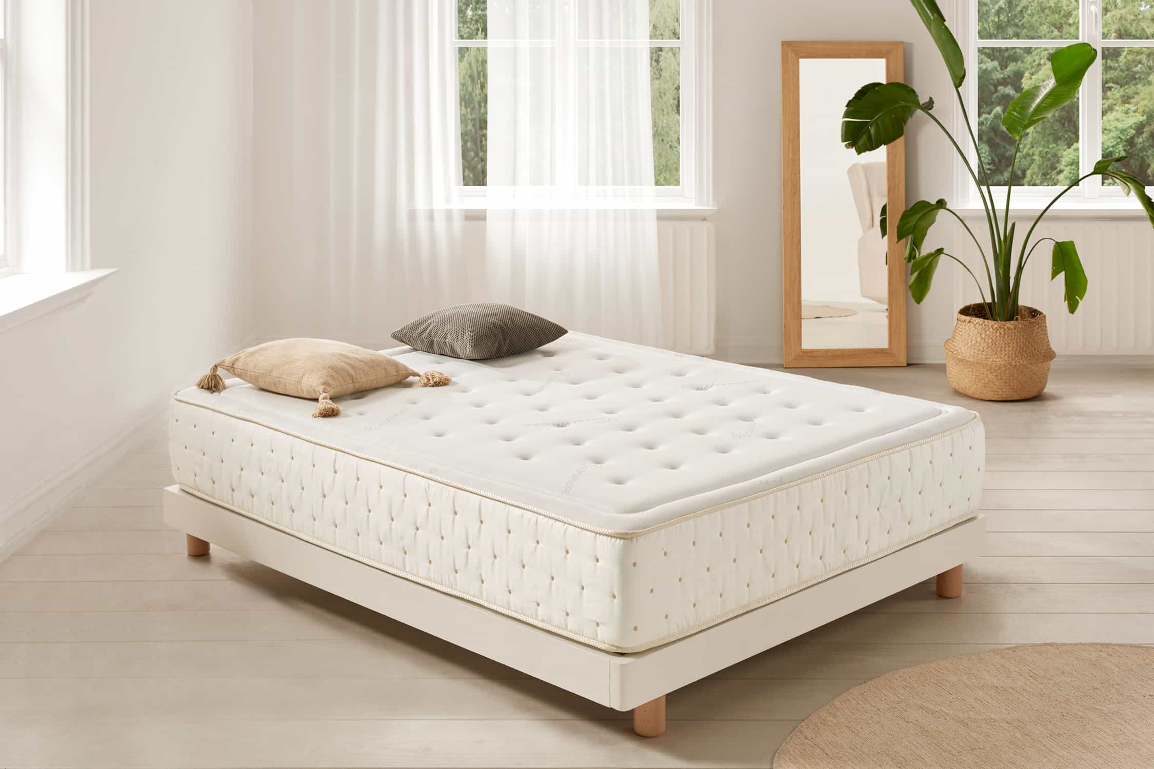Dormitorio con colchón sobre somier blanco. Hay almohadas, una planta y un espejo.