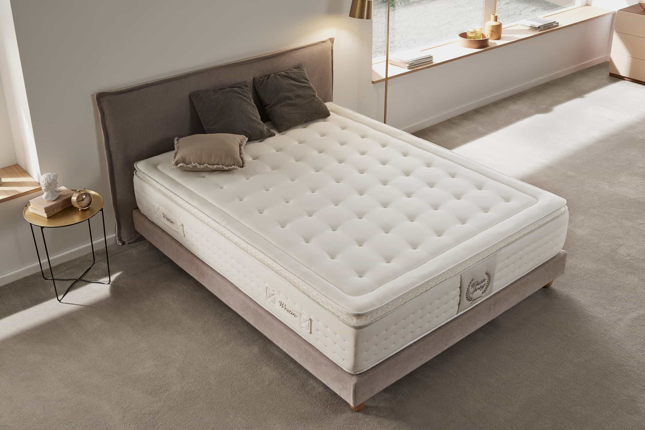Un colchón blanco está sobre una cama, con almohadas, una mesita de noche y una lámpara en la habitación.