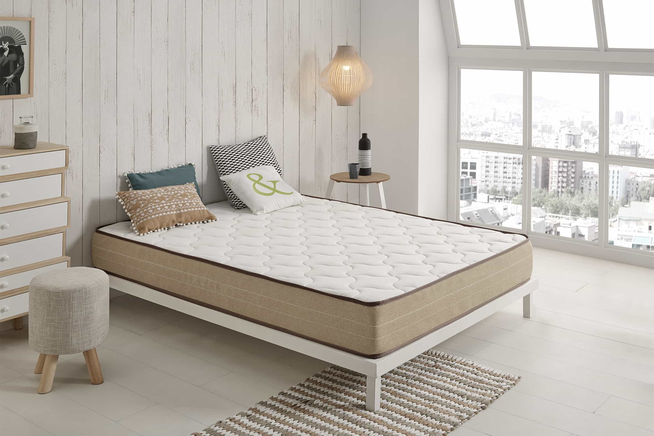Cama blanca con colchón y almohadas en una habitación luminosa. Hay una ventana al fondo.