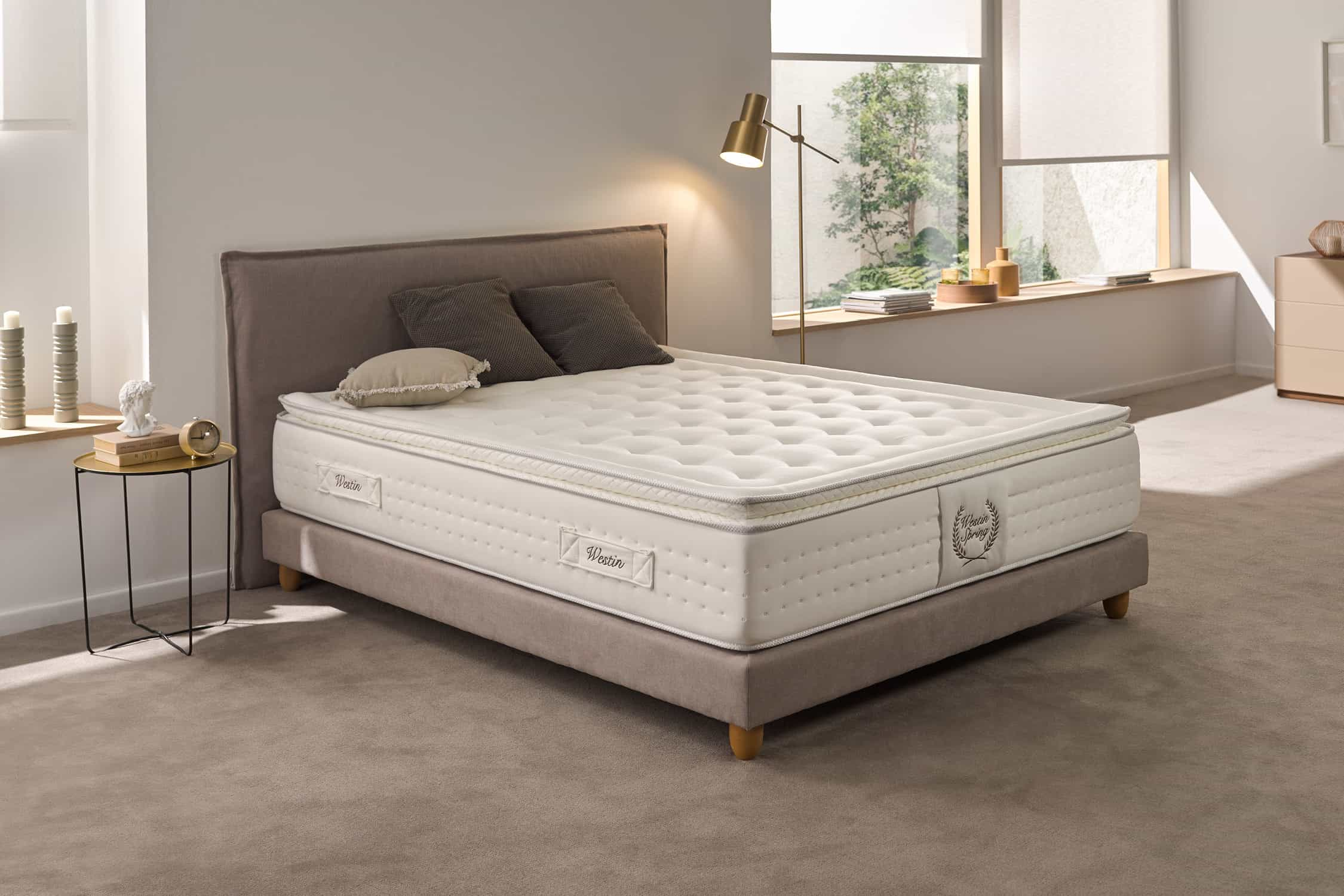 Una cama con un colchón blanco. Dos almohadas y una manta están en la cama. La habitación tiene una paleta de colores neutros.