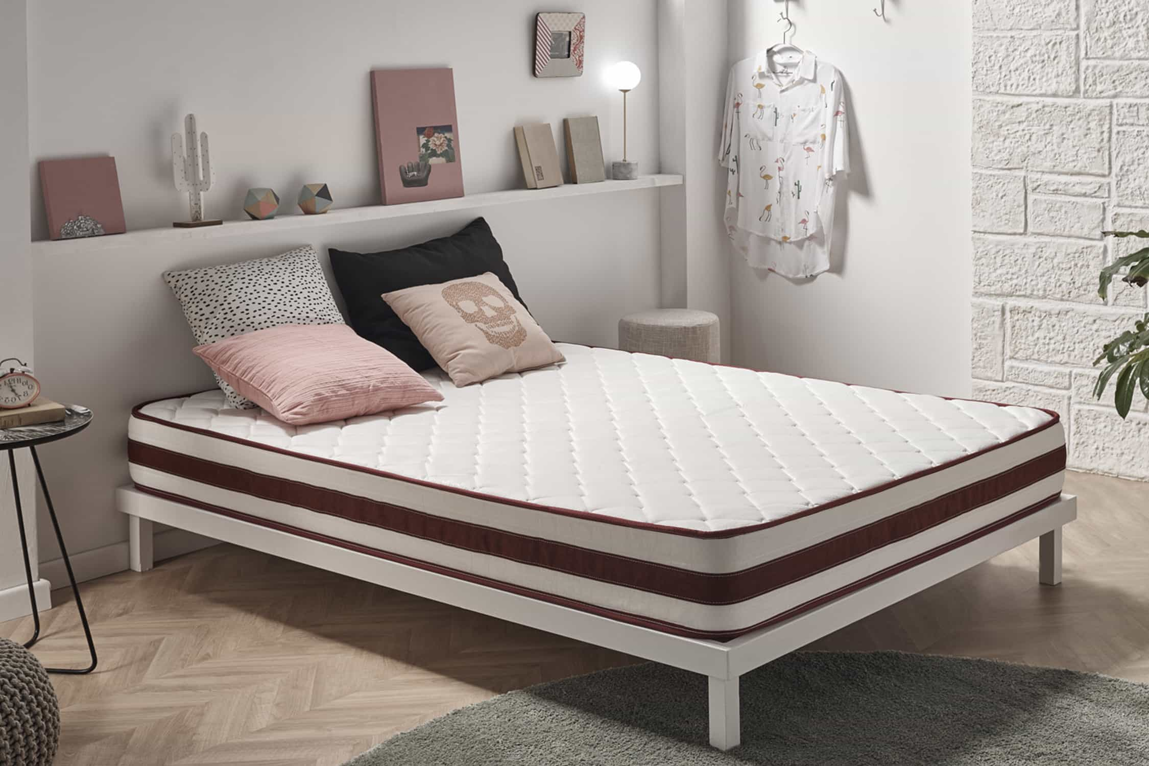 Cama blanca con colchón blanco y ribete rojo. Almohadas y decoración sobre fondo de pared blanca.