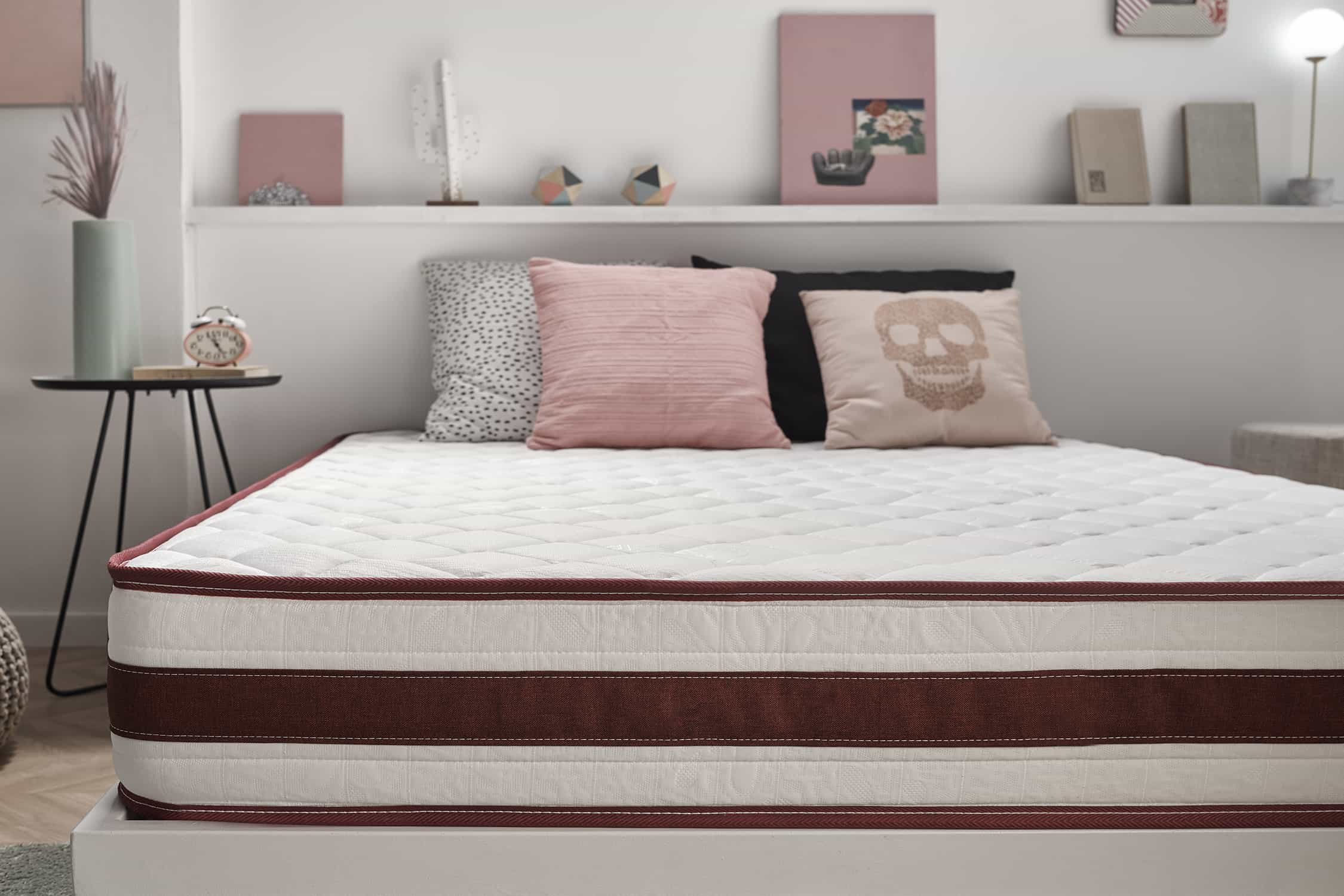 Una cama con colchón blanco y borde granate. Hay almohadas en la cama. Estantes y decoración al fondo.