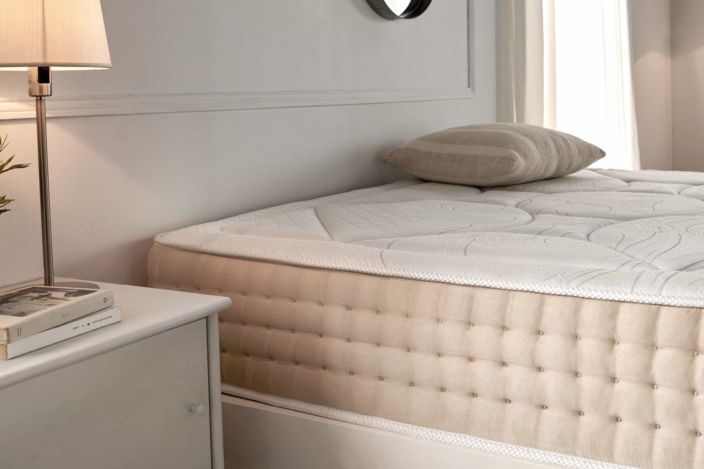 Una cama con un colchón blanco. Hay una almohada en la cama. Lámpara y mesita de noche a la izquierda. Paredes blancas.