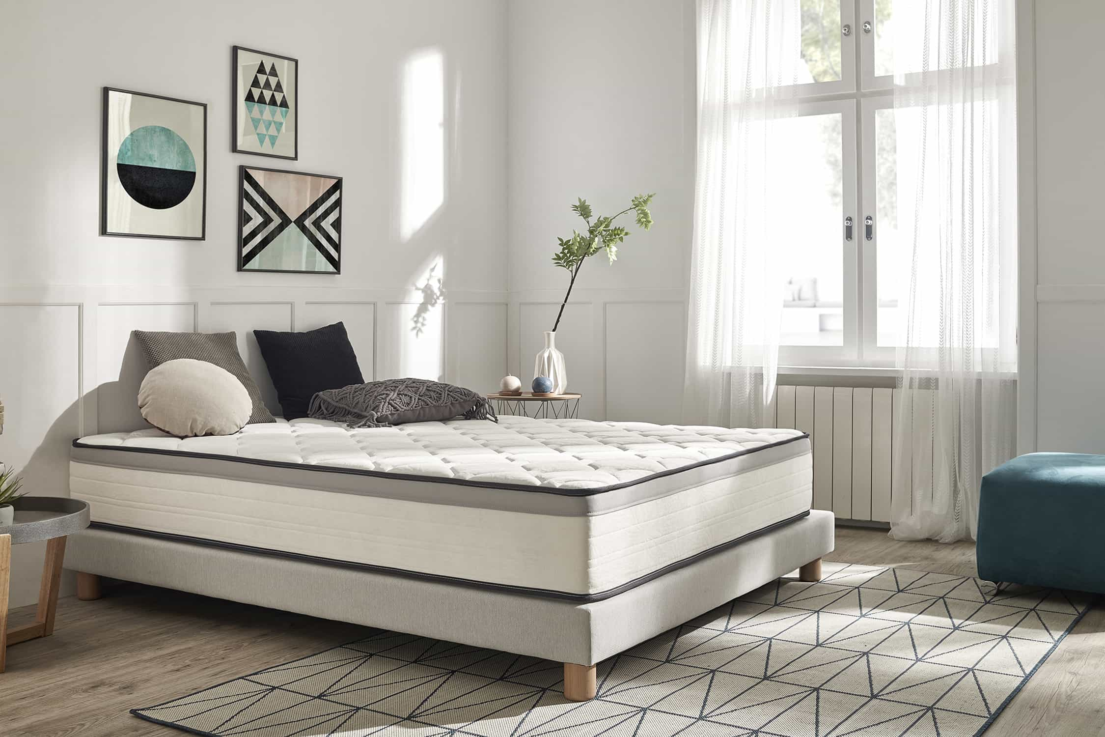 Una cama con colchón blanco y borde gris. Hay almohadas en la cama. Obras de arte y muebles al fondo.