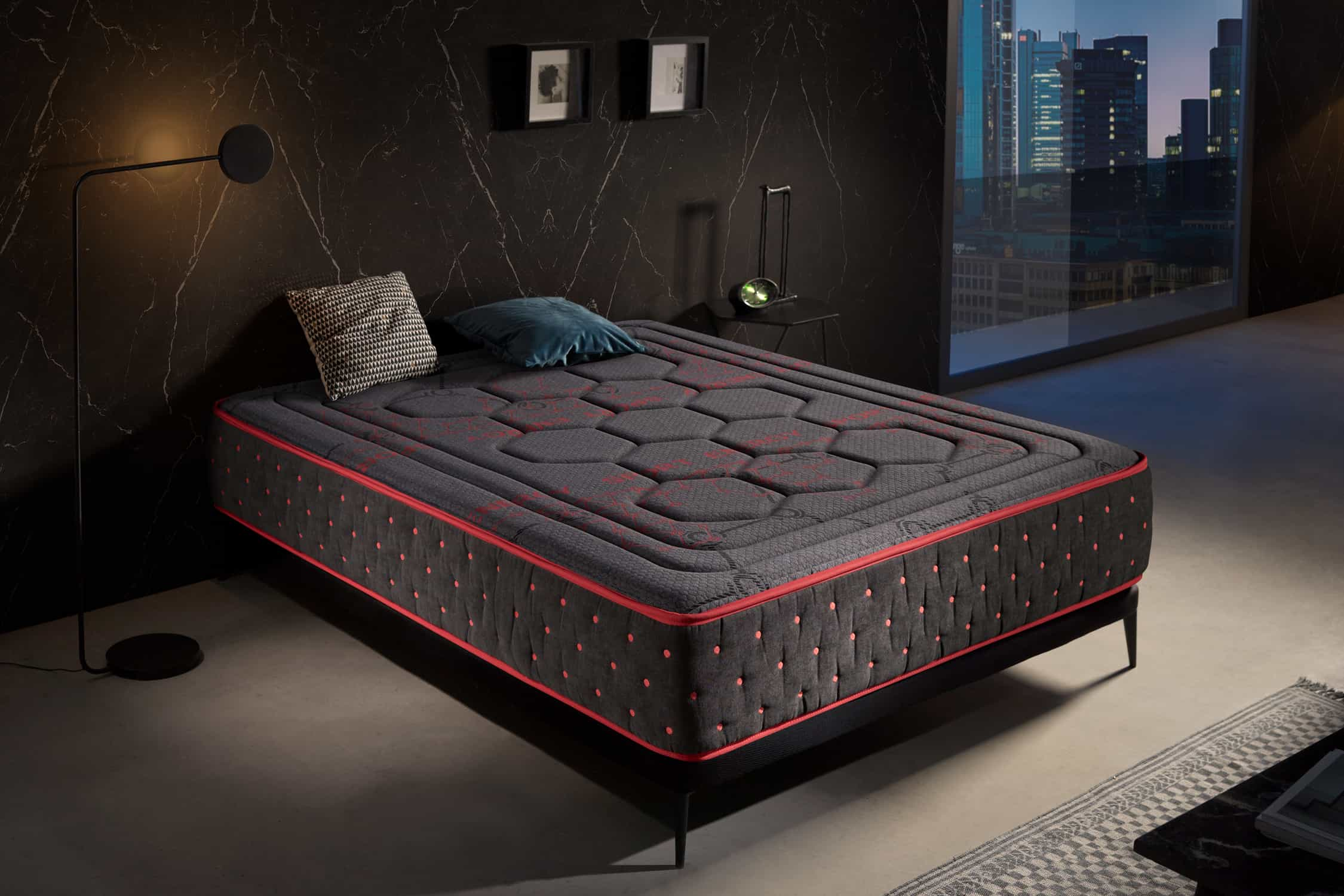 Una cama con un colchón gris y rojo estampado. Hay una almohada en la cama. La habitación tiene paredes y decoración oscuras.