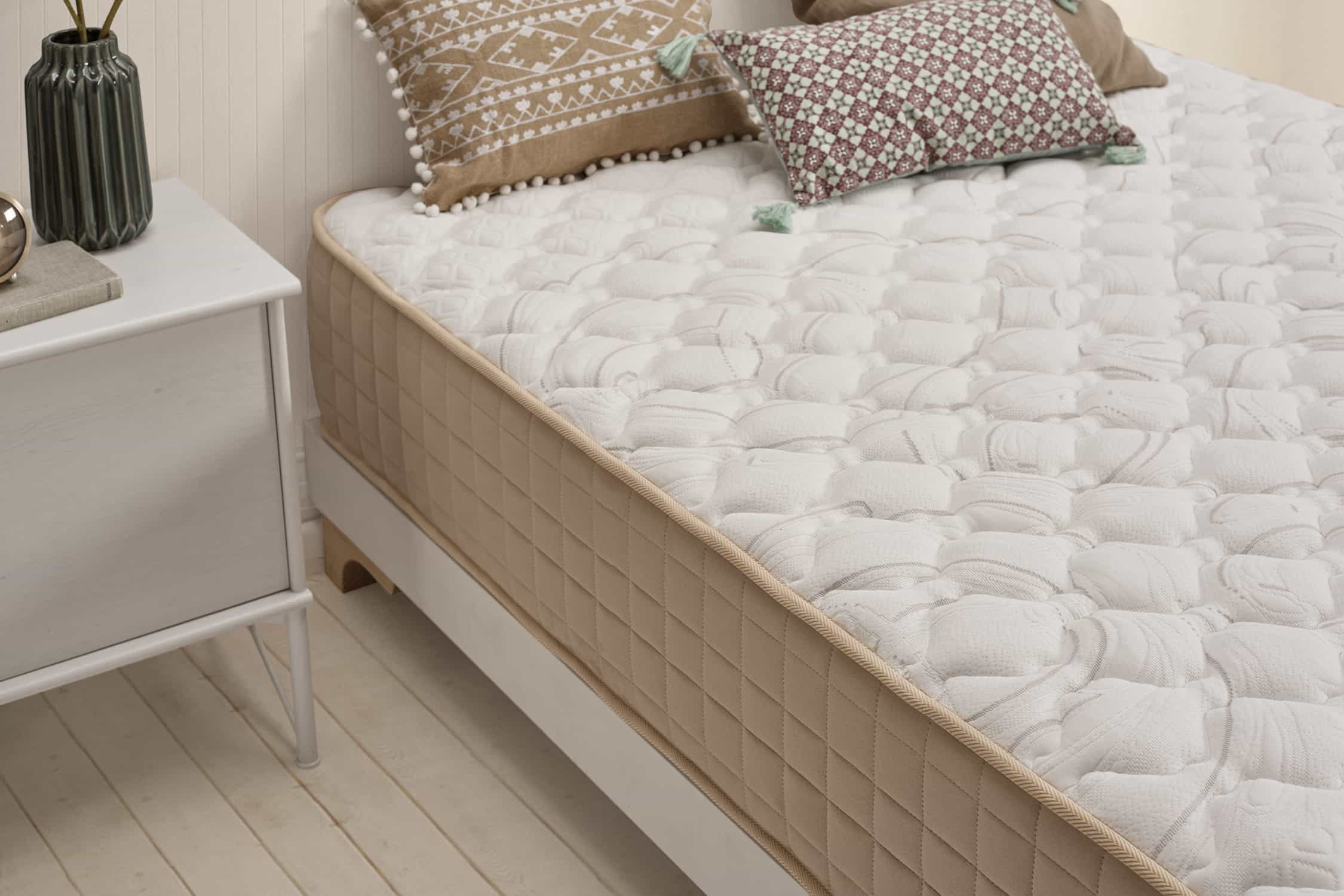 Una cama con colchón blanco, almohadas y una mesita de noche blanca. Un pequeño jarrón en la mesa.