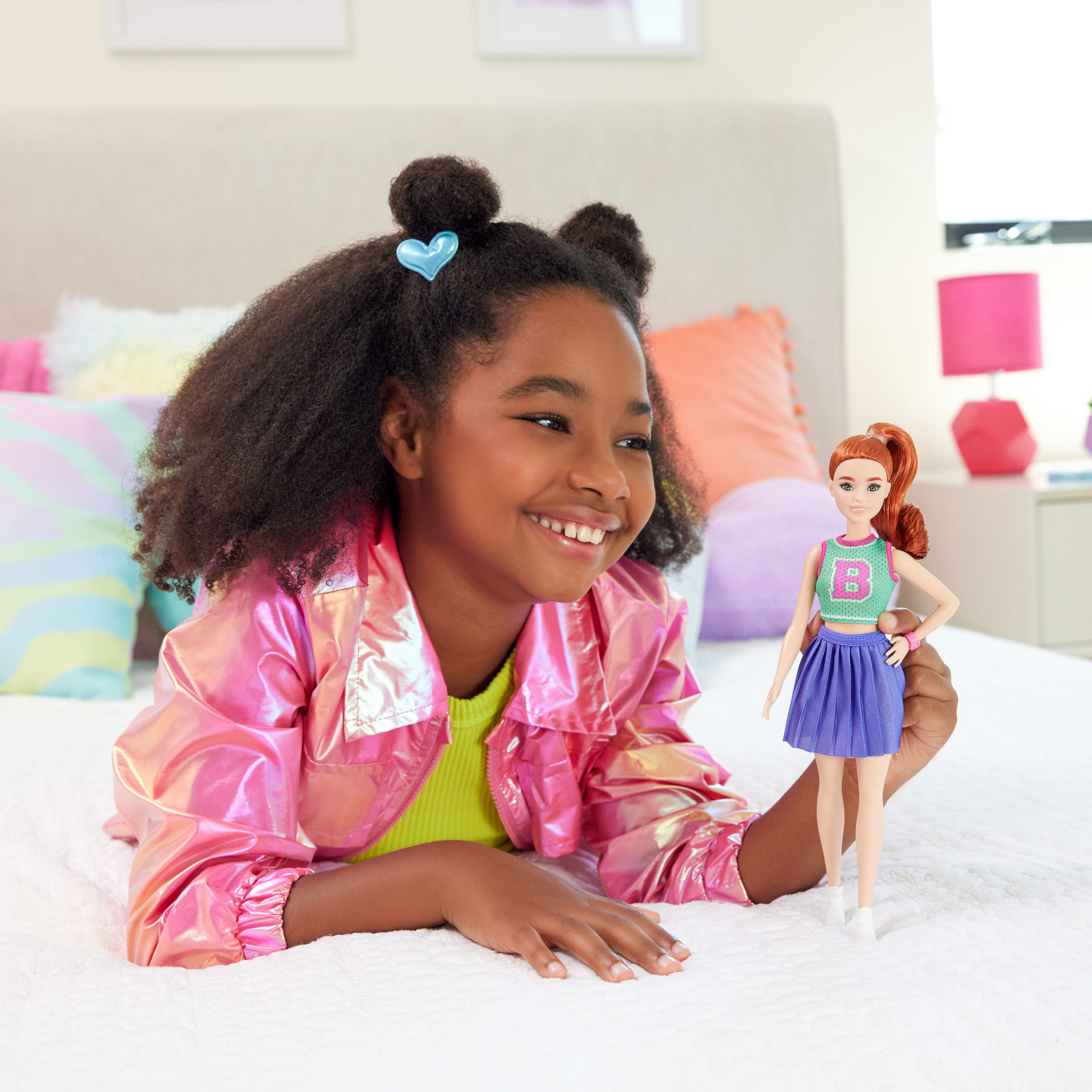 Dziewczyna z kręconymi włosami leży na łóżku z lalką. Ma na sobie różową kurtkę i uśmiecha się.