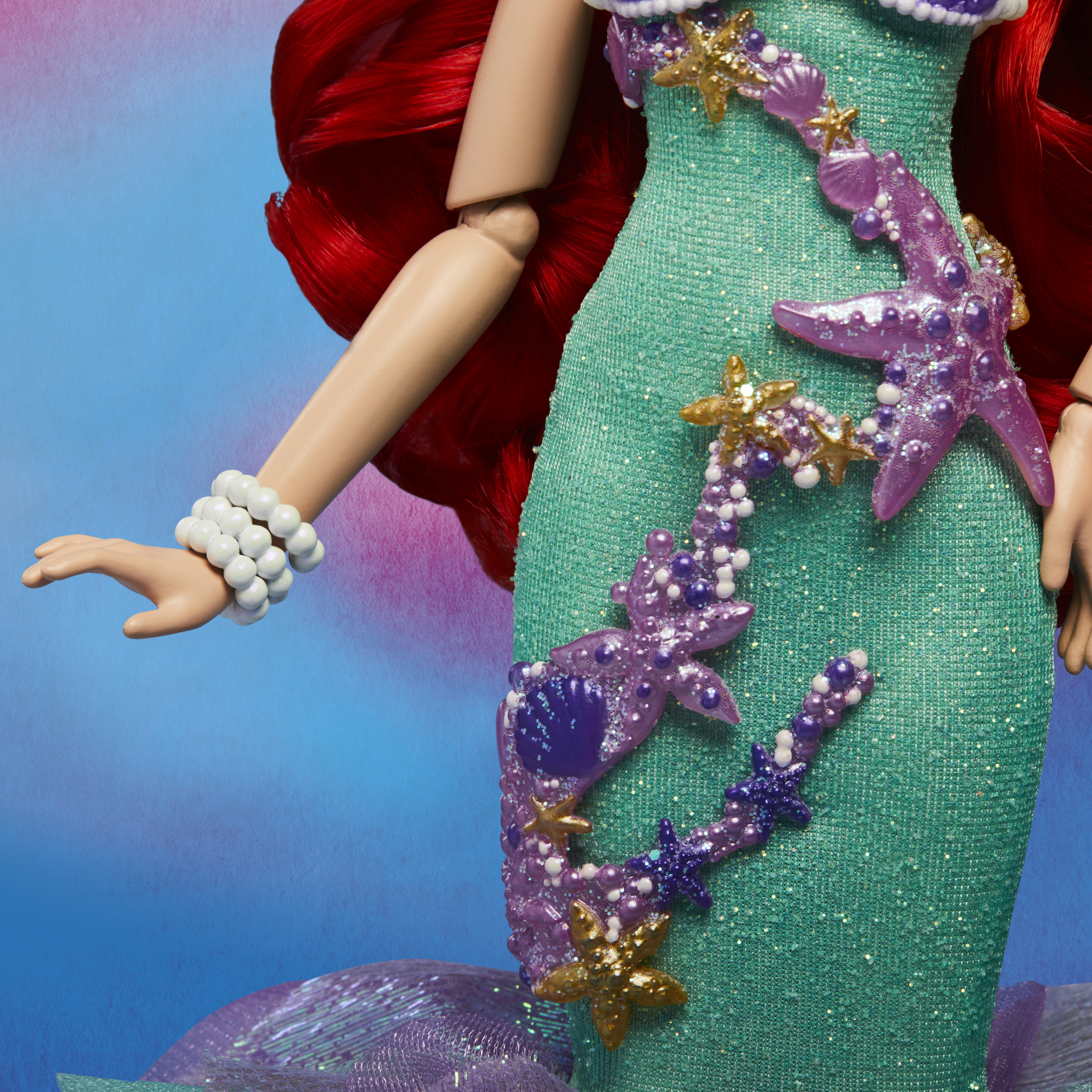 Nahaufnahme des Oberkörpers einer Meerjungfrauenpuppe. Sie hat rotes Haar, ein grünes Kleid mit sternförmigen Verzierungen und ein weißes Armband.