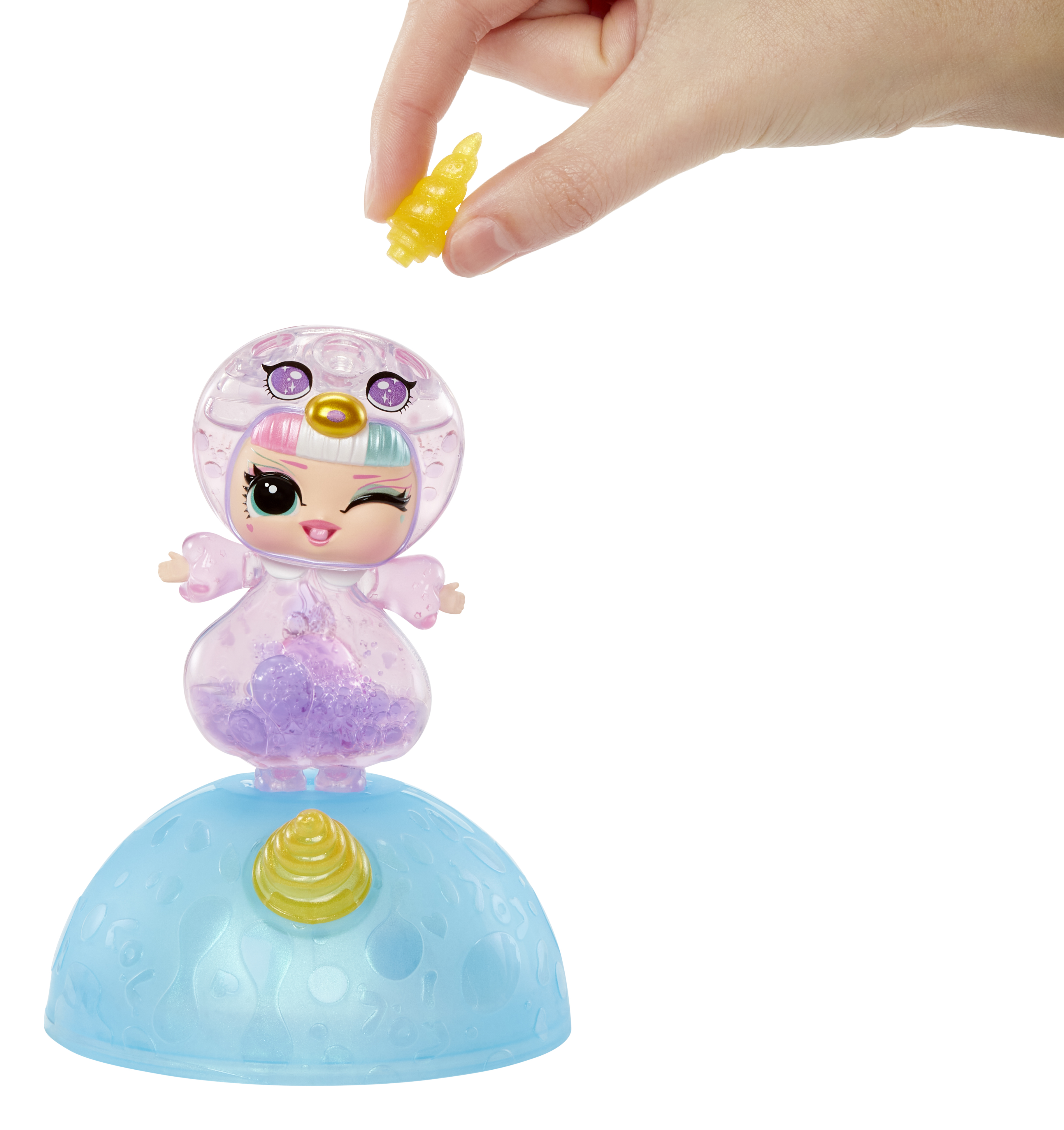 Una mano coloca un cuerno amarillo sobre una muñeca en una cúpula transparente. La muñeca es rosa y morada.