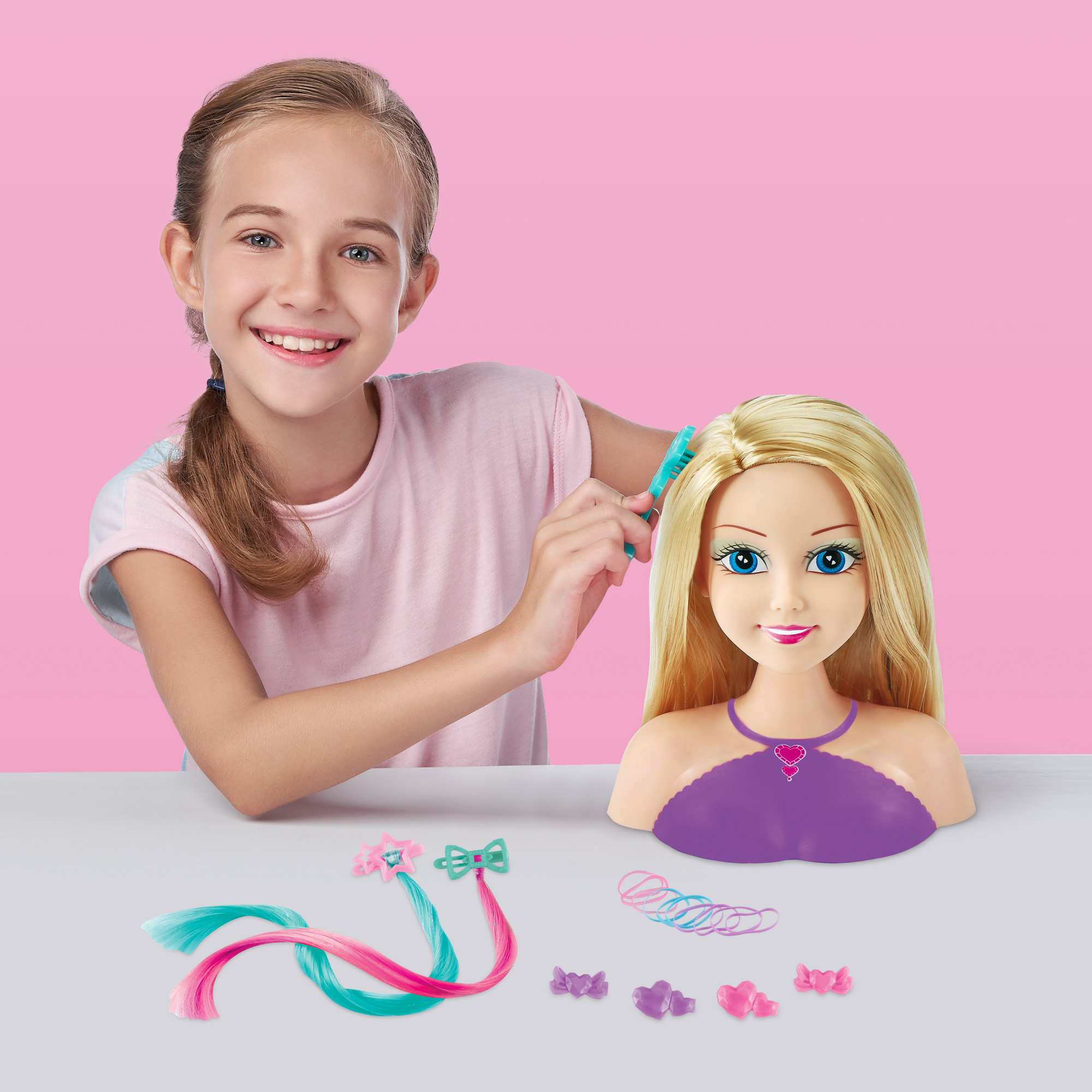 Dziewczyna czesze blond włosy lalki. Głowa lalki na stole z akcesoriami do włosów. Różowe tło.