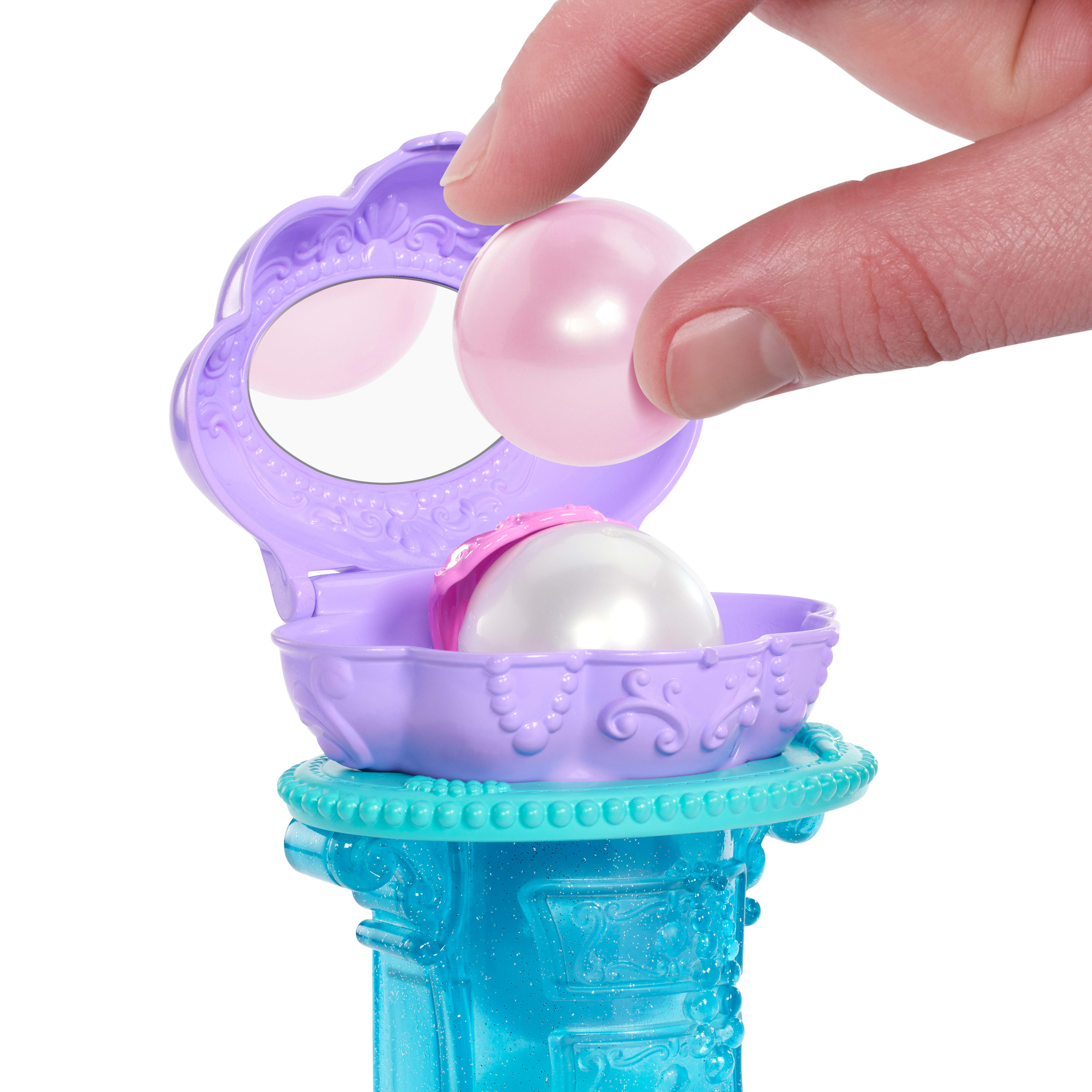 Ręka umieszcza różową perłę w fioletowym etui w kształcie muszli z lustrem. Niebieska podstawa.