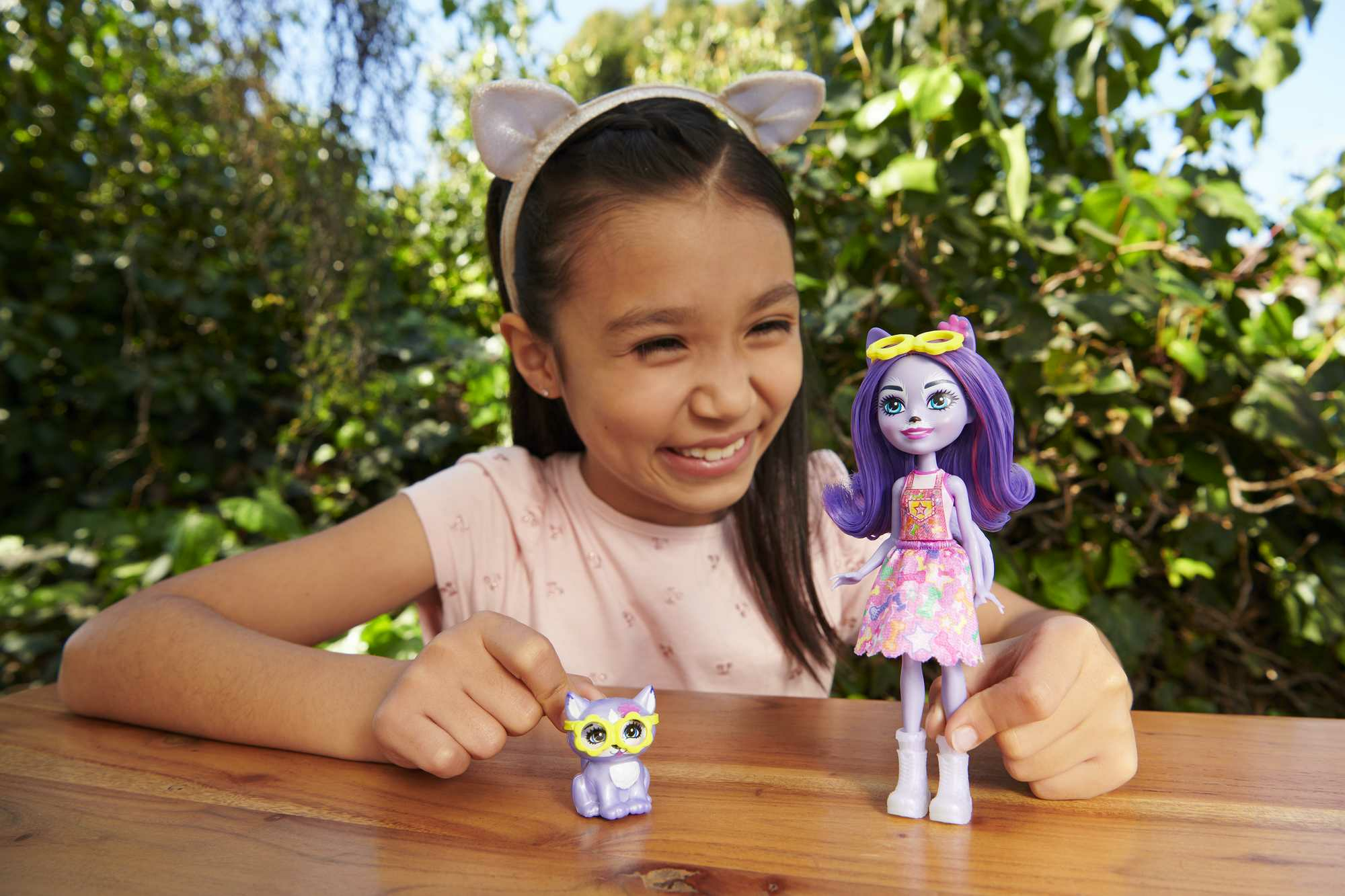 Mädchen mit Puppe und Spielzeugkatze. Das Mädchen lächelt. Hintergrund ist ein Garten mit grünen Pflanzen.