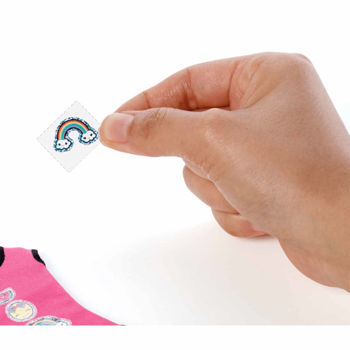 Eine Hand hält einen kleinen Regenbogen-Aufkleber über einem rosa Hemd mit anderen Aufklebern.