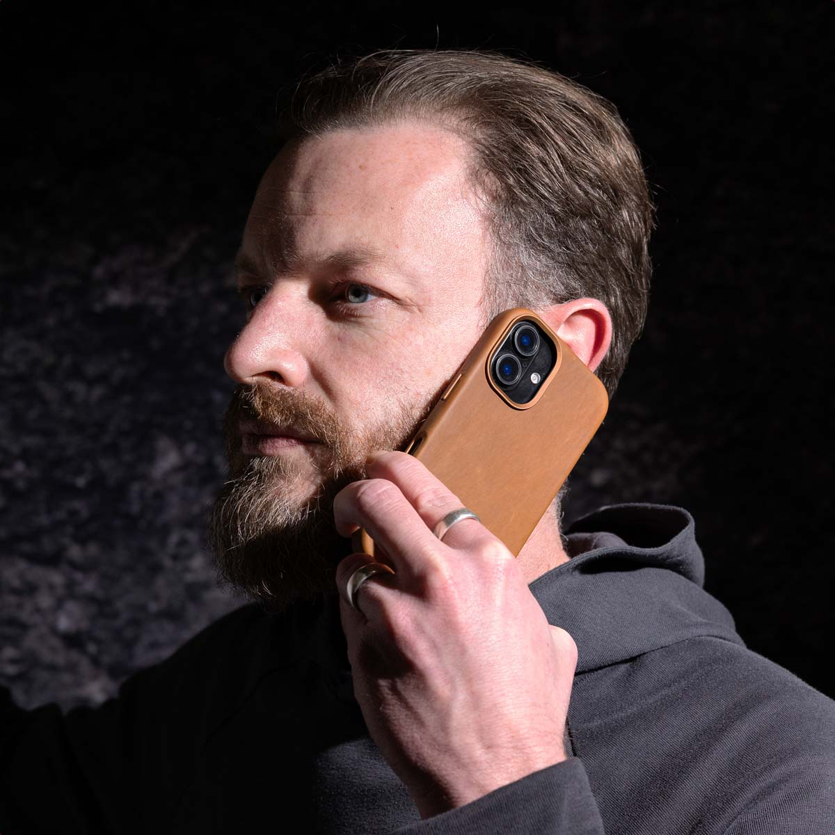 Homme barbu utilisant un étui de téléphone marron. Fond sombre. Bague au doigt.