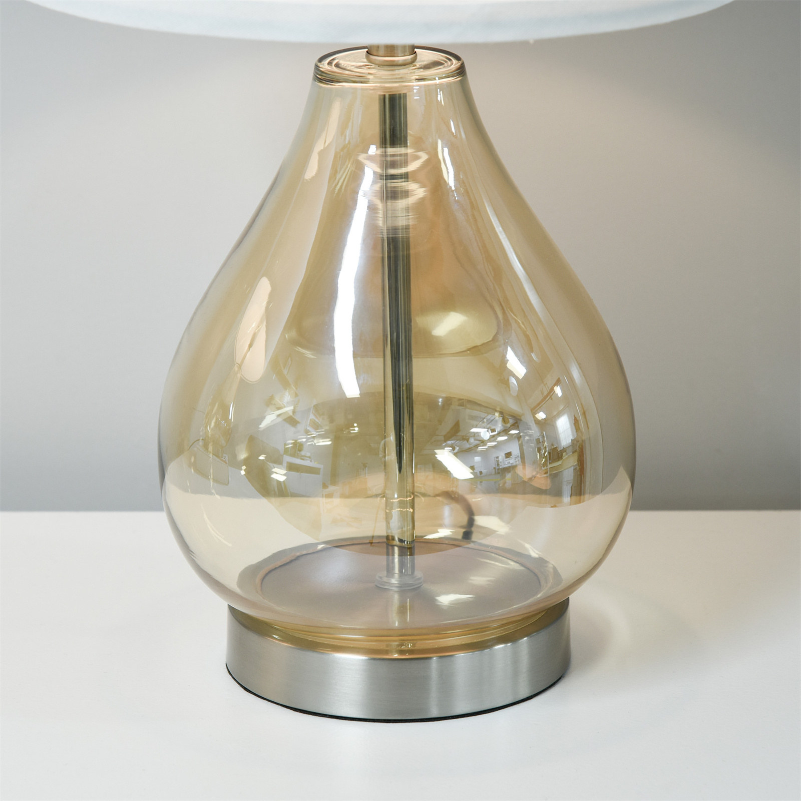 Eine Tischlampe mit tropfenförmigem, bernsteinfarbenem Glasfuß und weißem Lampenschirm auf weißer Oberfläche.