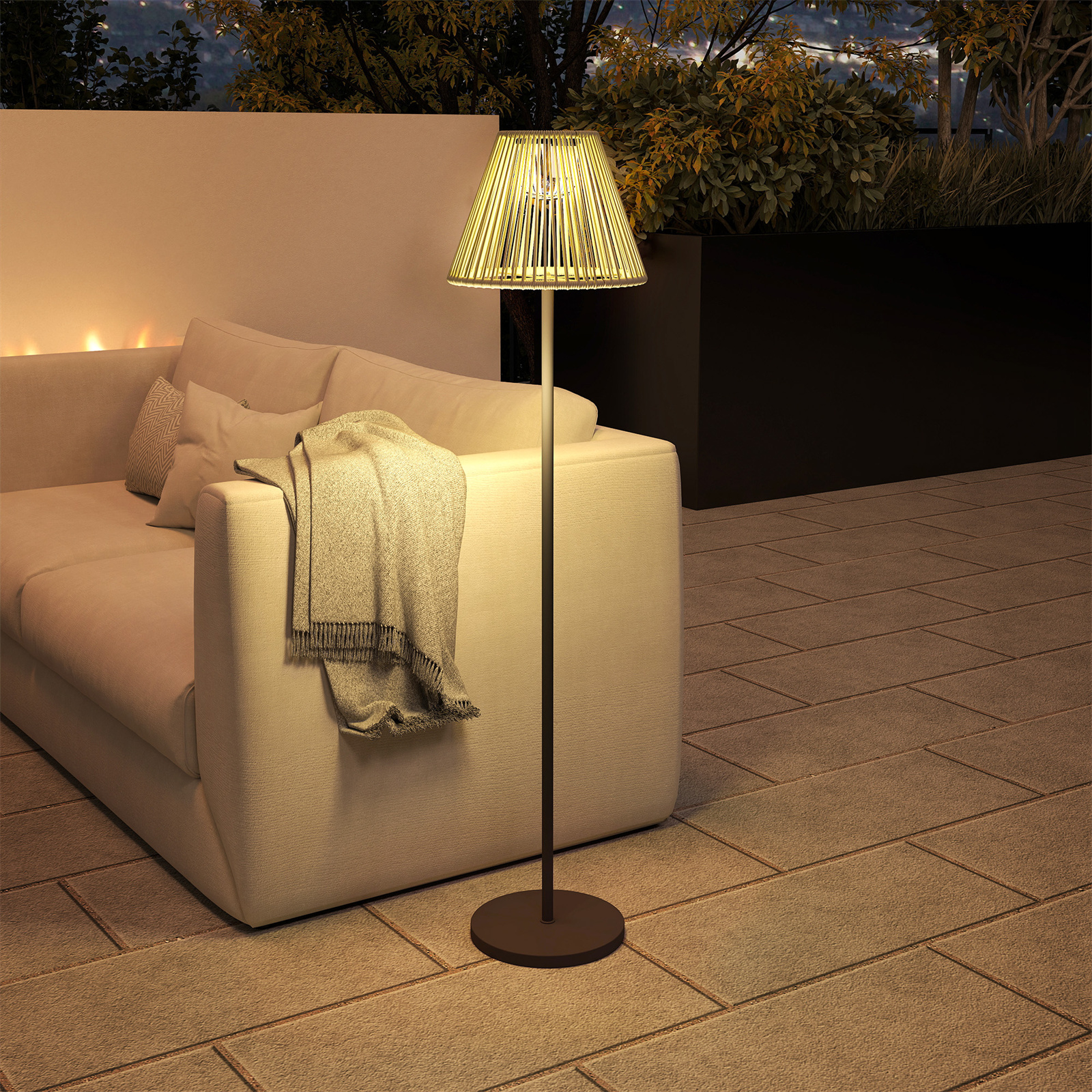 Eine Stehlampe mit gewebtem Lampenschirm neben einem Sofa und einer Decke auf einer Terrasse in der Dämmerung.