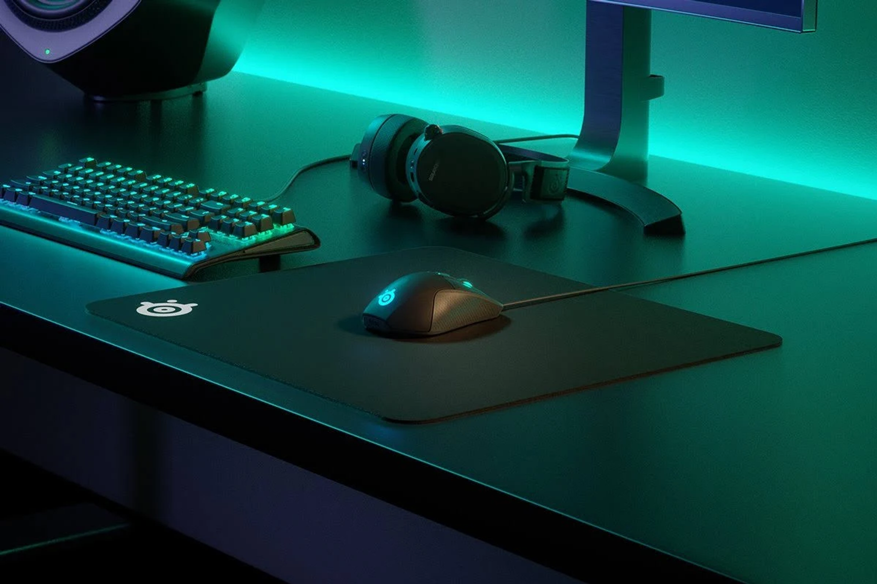 Gaming-Setup mit Tastatur, Maus, Mauspad, Kopfhörern und Monitor. Grüne und blaue Beleuchtung.