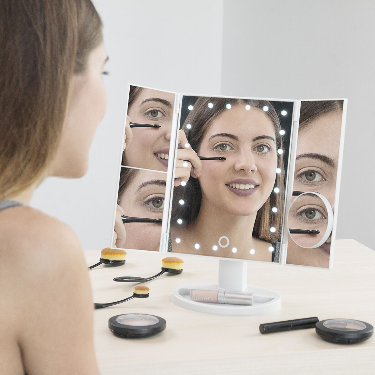 Mujer usando espejo de maquillaje triple iluminado, con brochas y maquillaje.