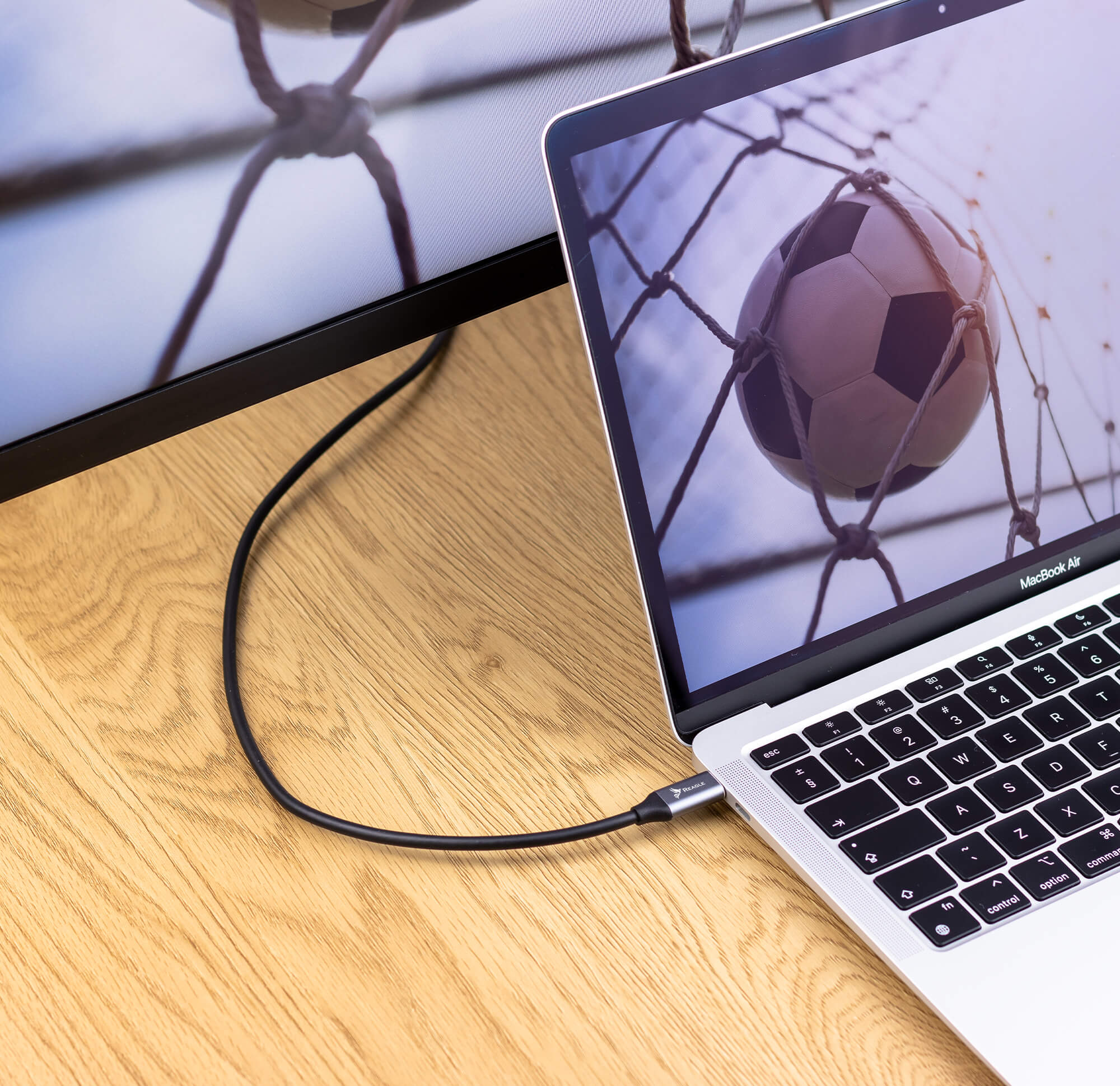 Laptop podłączony do monitora pokazującego piłkę nożną w siatce, na drewnianym stole.