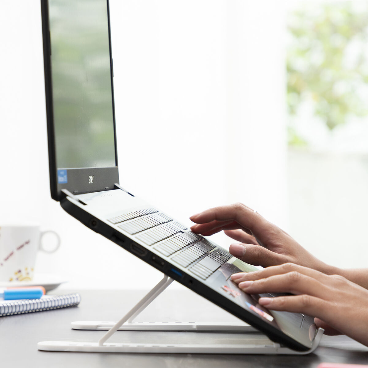 Persona che digita su un laptop su un supporto bianco. Una tazza di caffè e una penna sono sul tavolo.