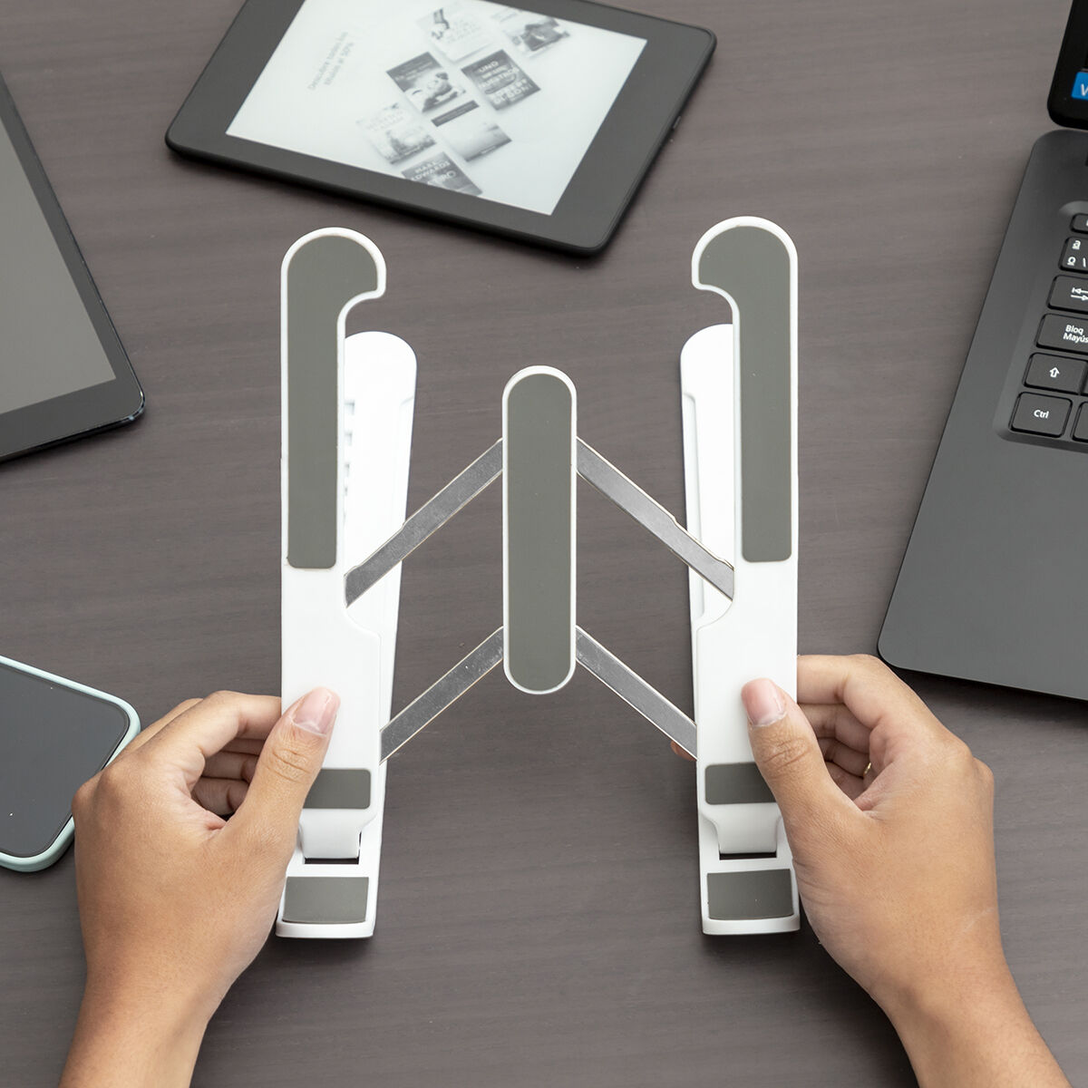 Mani che tengono un supporto per laptop bianco e grigio. C'è un tablet e un telefono sulla scrivania.