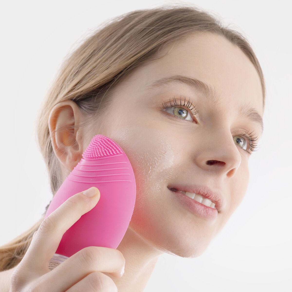 Donna che usa una spazzola rosa per la pulizia del viso sul viso, sorridendo. Sfondo bianco, primo piano.