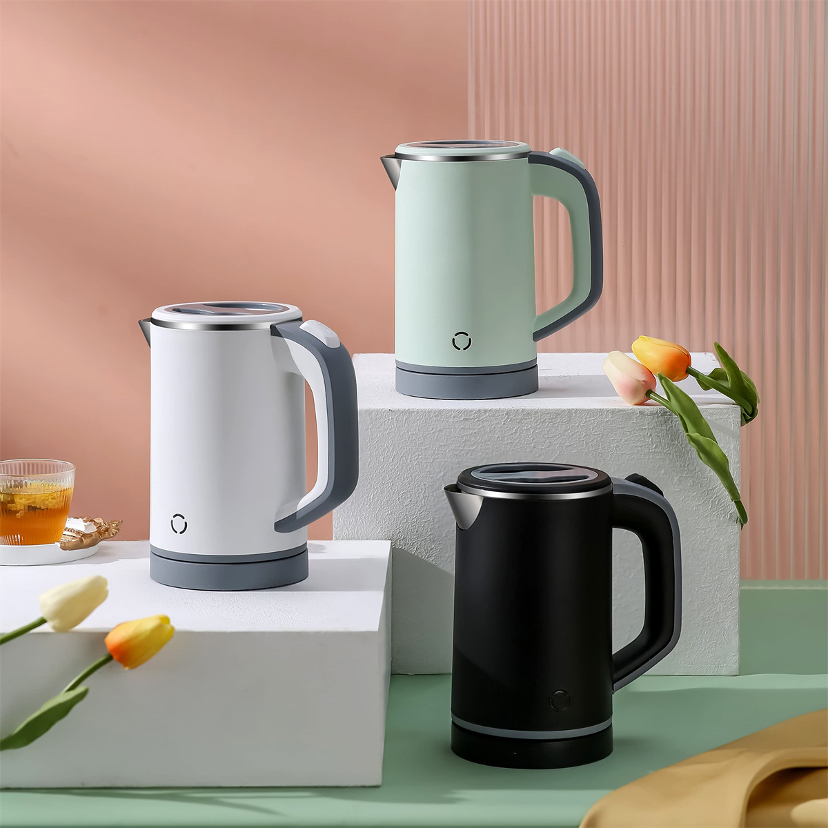 Drei elektrische Wasserkocher in Weiß, Grün und Schwarz. Sie stehen auf einer weißen Oberfläche. Blumen und eine Tasse Tee sind in der Nähe.