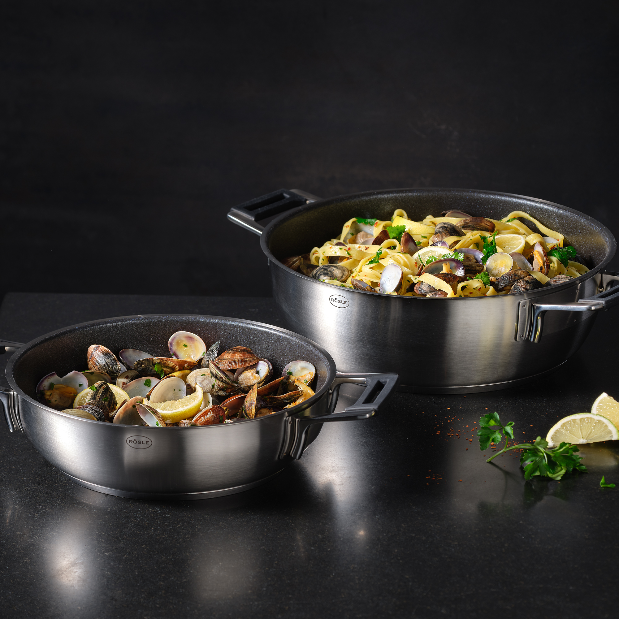 Zwei Edelstahlpfannen mit Essen. Eine mit Muscheln, eine mit Pasta.