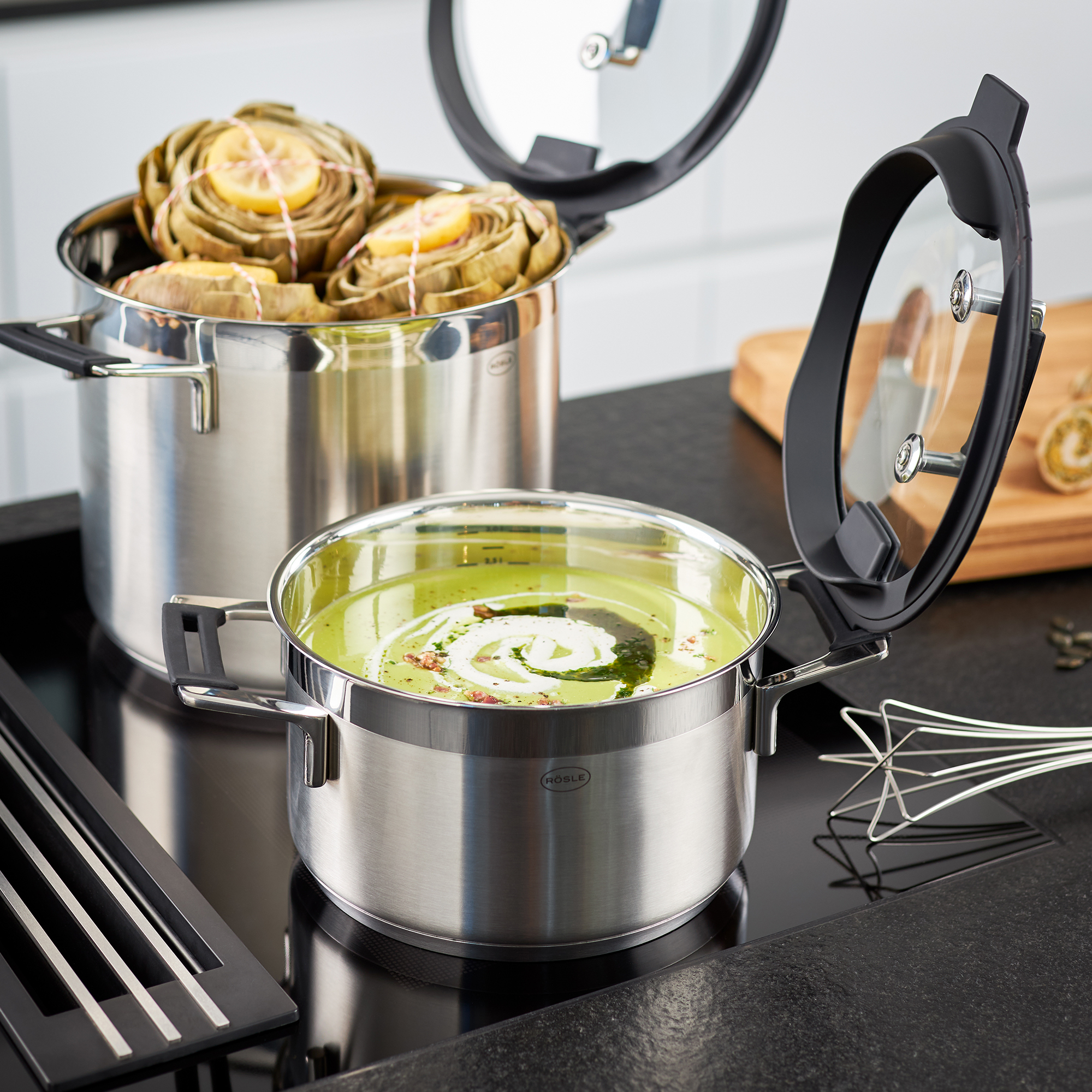 Deux casseroles en acier inoxydable avec de la nourriture. L'une a de la soupe, l'autre des artichauts. Comptoir noir.