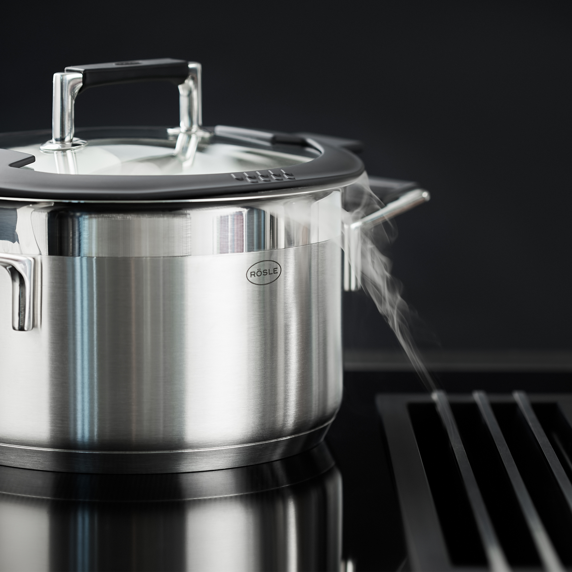 Pot en acier inoxydable avec de la vapeur qui monte. Le couvercle est dessus. Surface de cuisson noire.
