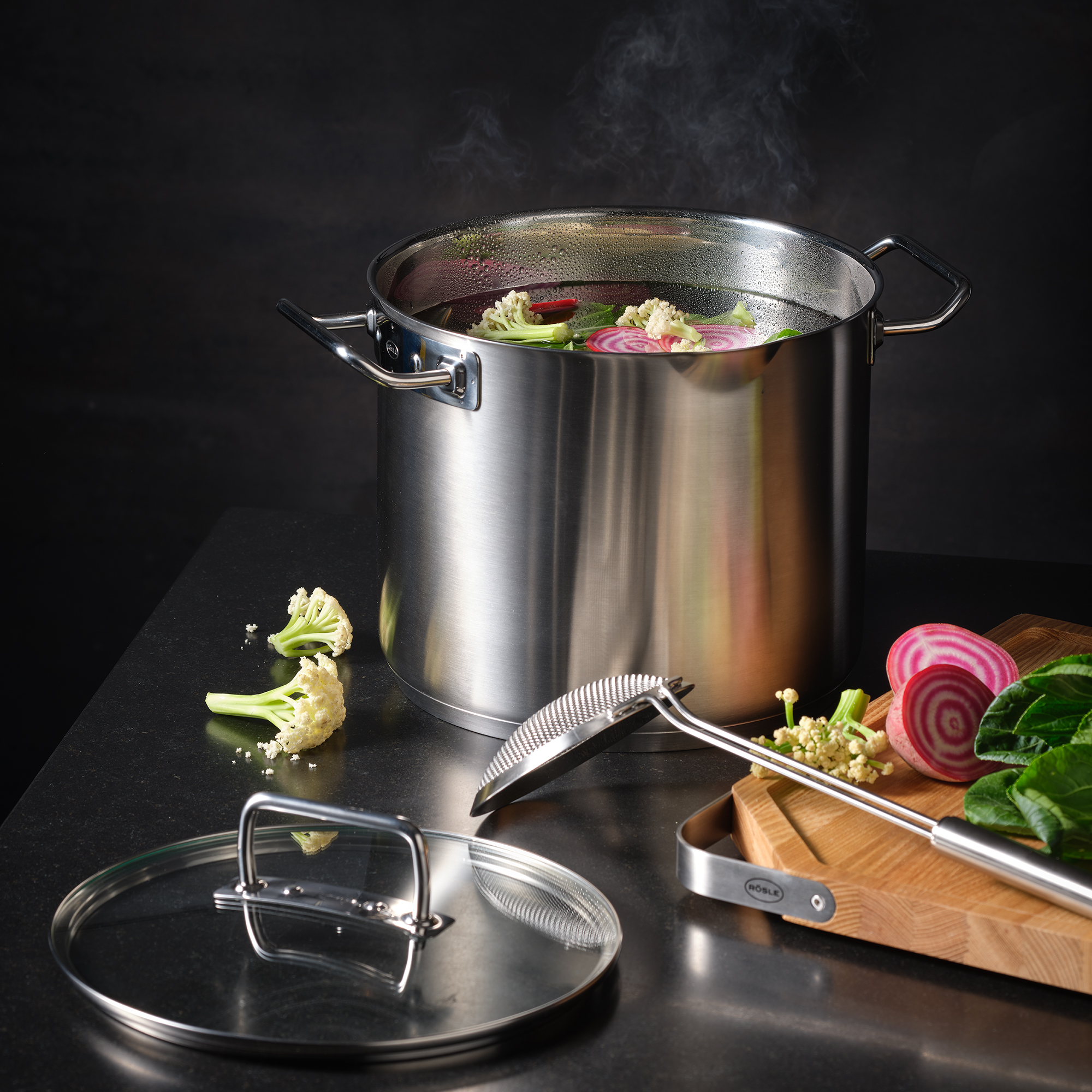 Grande casserole en acier inoxydable avec des légumes. Vapeur montante. Surface noire, planche de bois.
