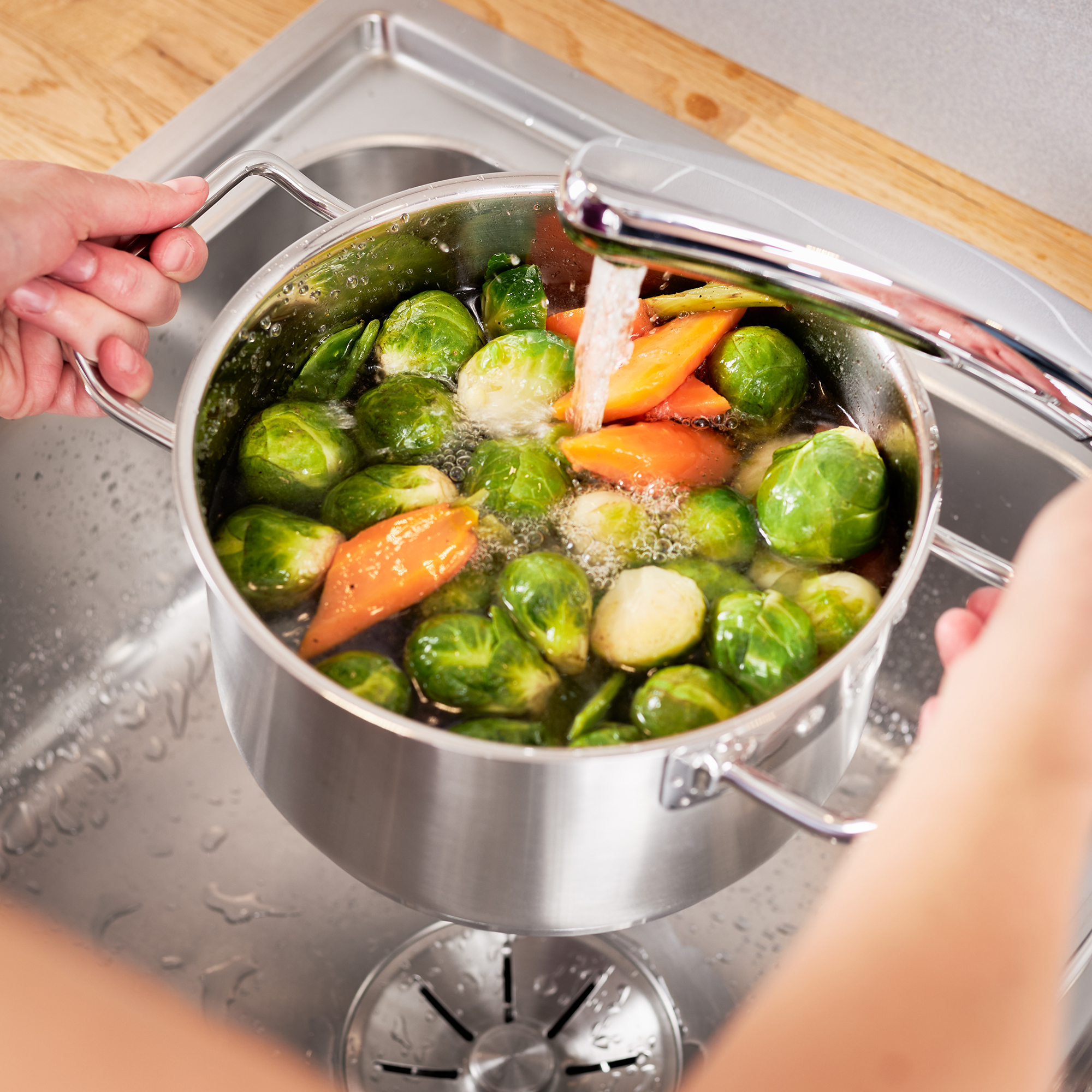 Une casserole en acier inoxydable remplie de choux de Bruxelles et de carottes sous un jet d'eau dans un évier.