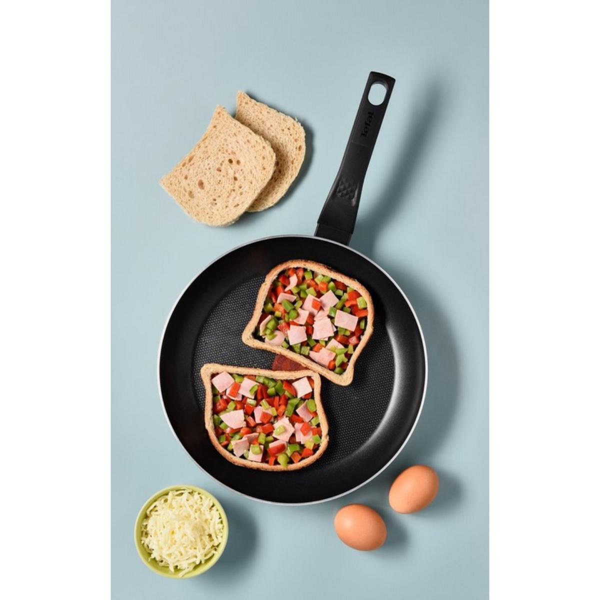 Frying pan with two toast slices topped with ham, veggies, and cheese. Blue background.