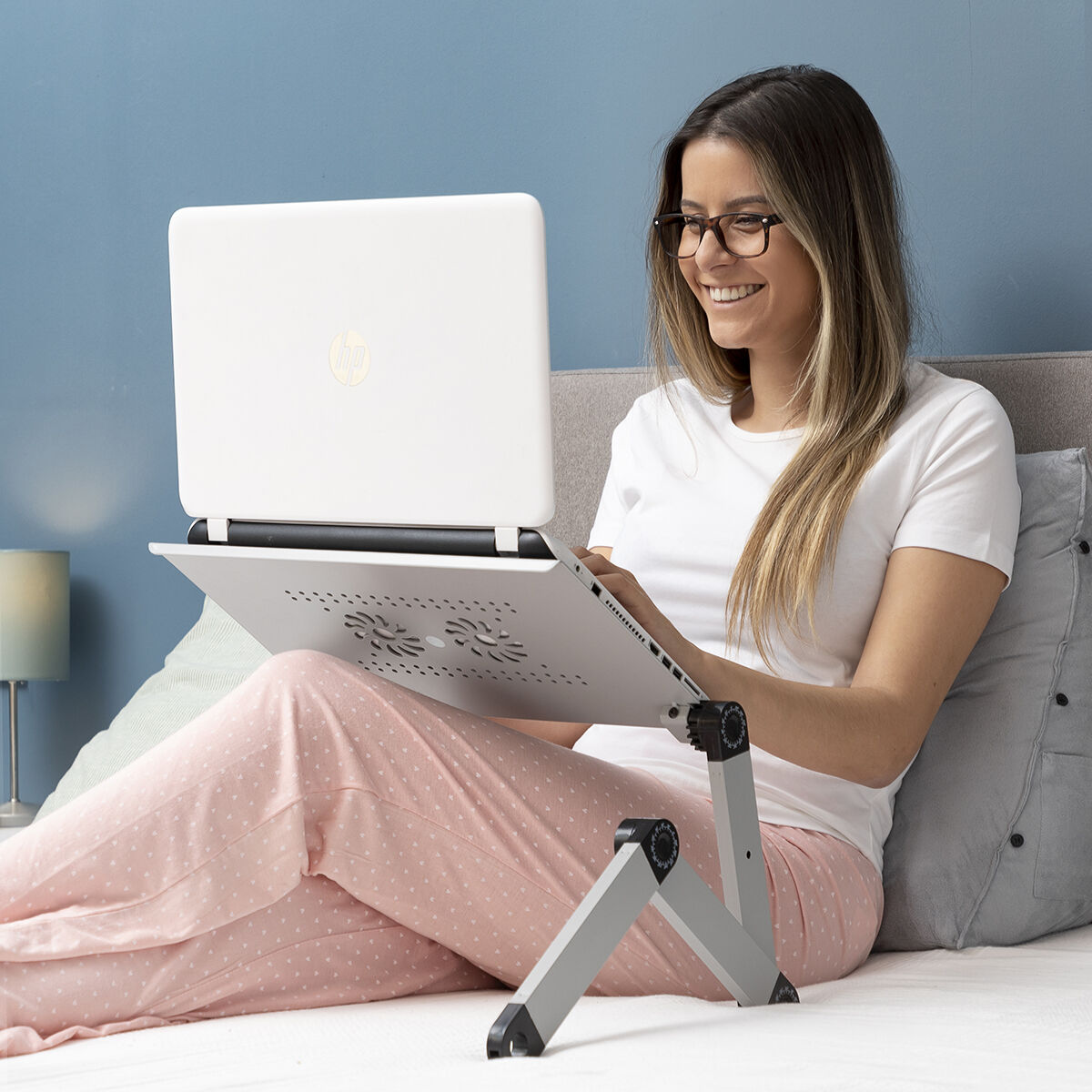 Una donna seduta su un letto con un laptop bianco su un supporto. Sta sorridendo.