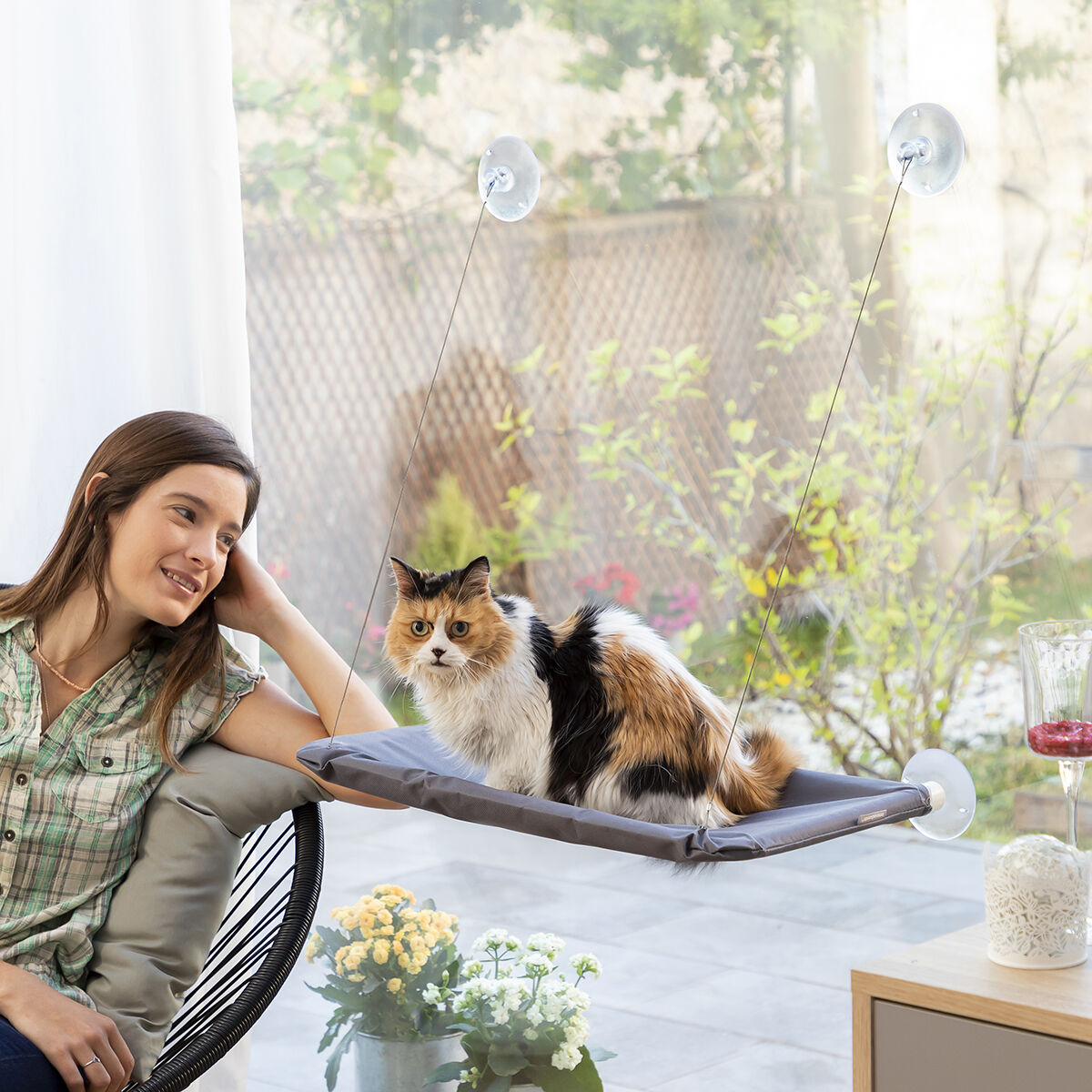 Mujer sentada, gato en una percha de ventana. La percha es gris, el gato tricolor. Ventana es fondo.