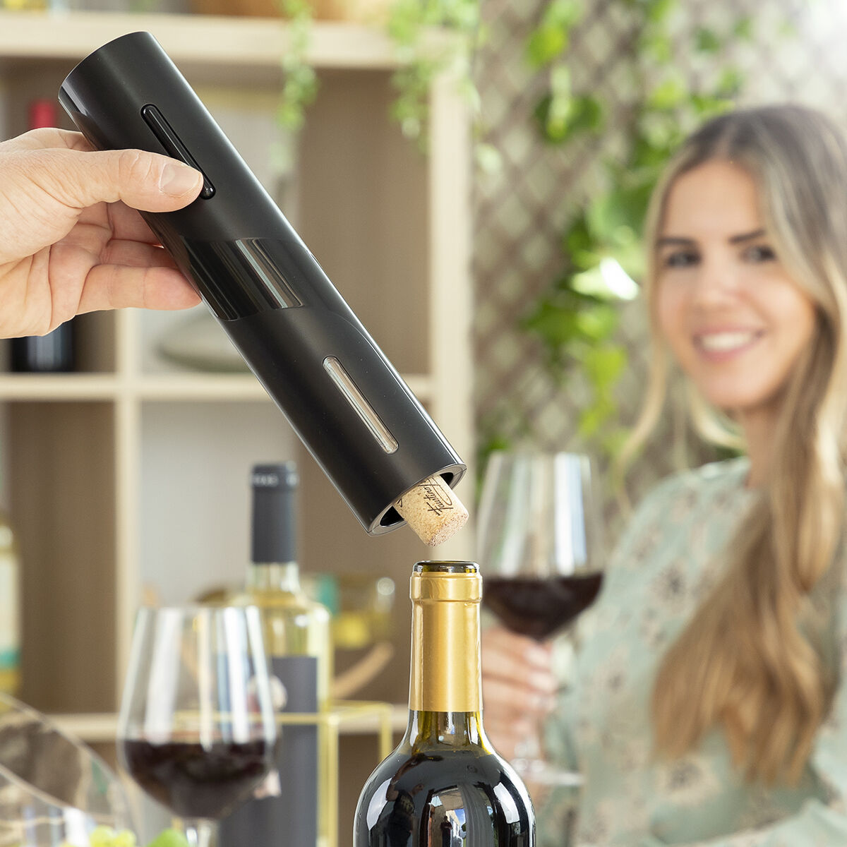 Una persona usa un abridor de vino eléctrico. Una botella y una mujer con copa de vino al fondo.