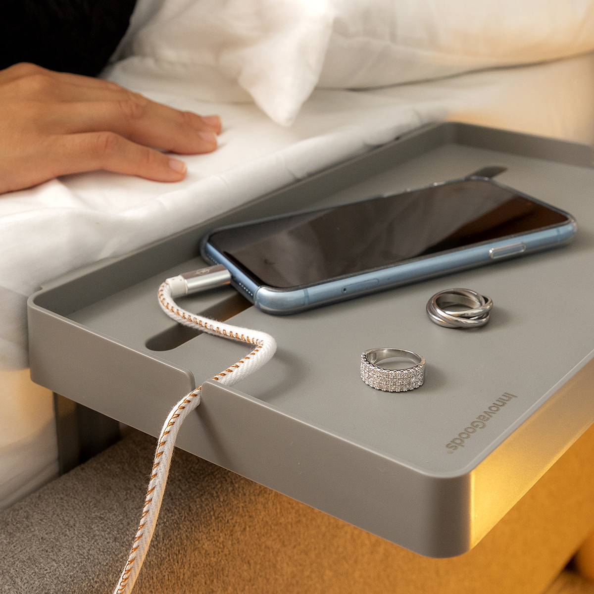 Vassoio grigio con telefono, anelli e cavo di ricarica. Letto bianco, luce gialla.