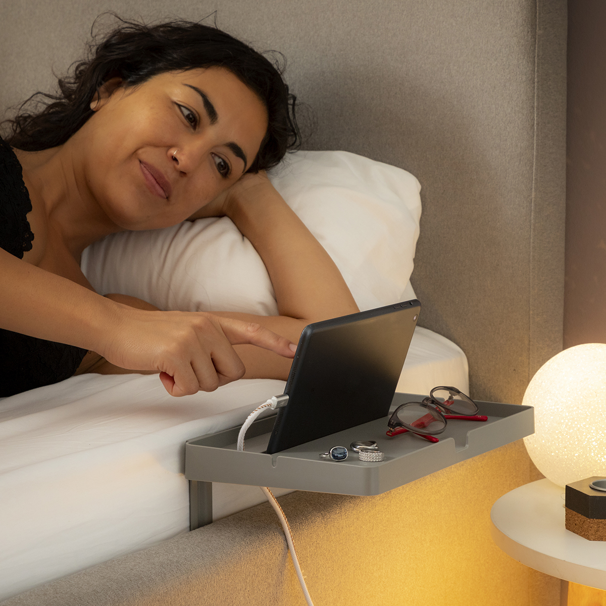 Donna a letto, usa un tablet. Occhiali e accessori su un vassoio. Lenzuola bianche, lampada.