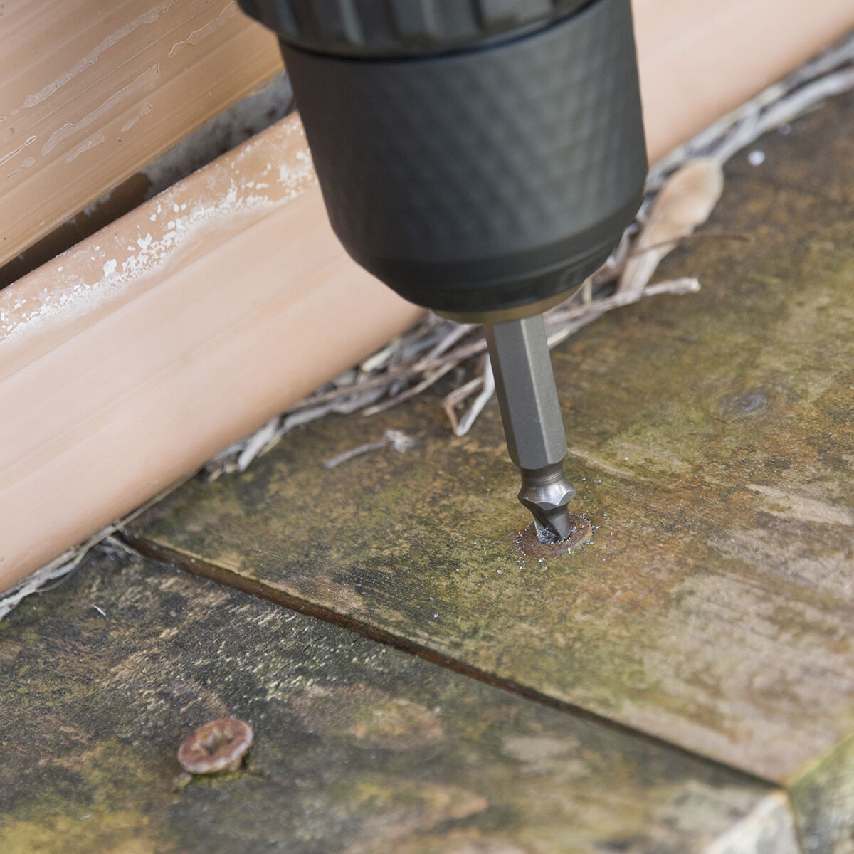 Una punta di trapano avvita una vite nel legno. Il legno è invecchiato e si vede un tubo arancione.