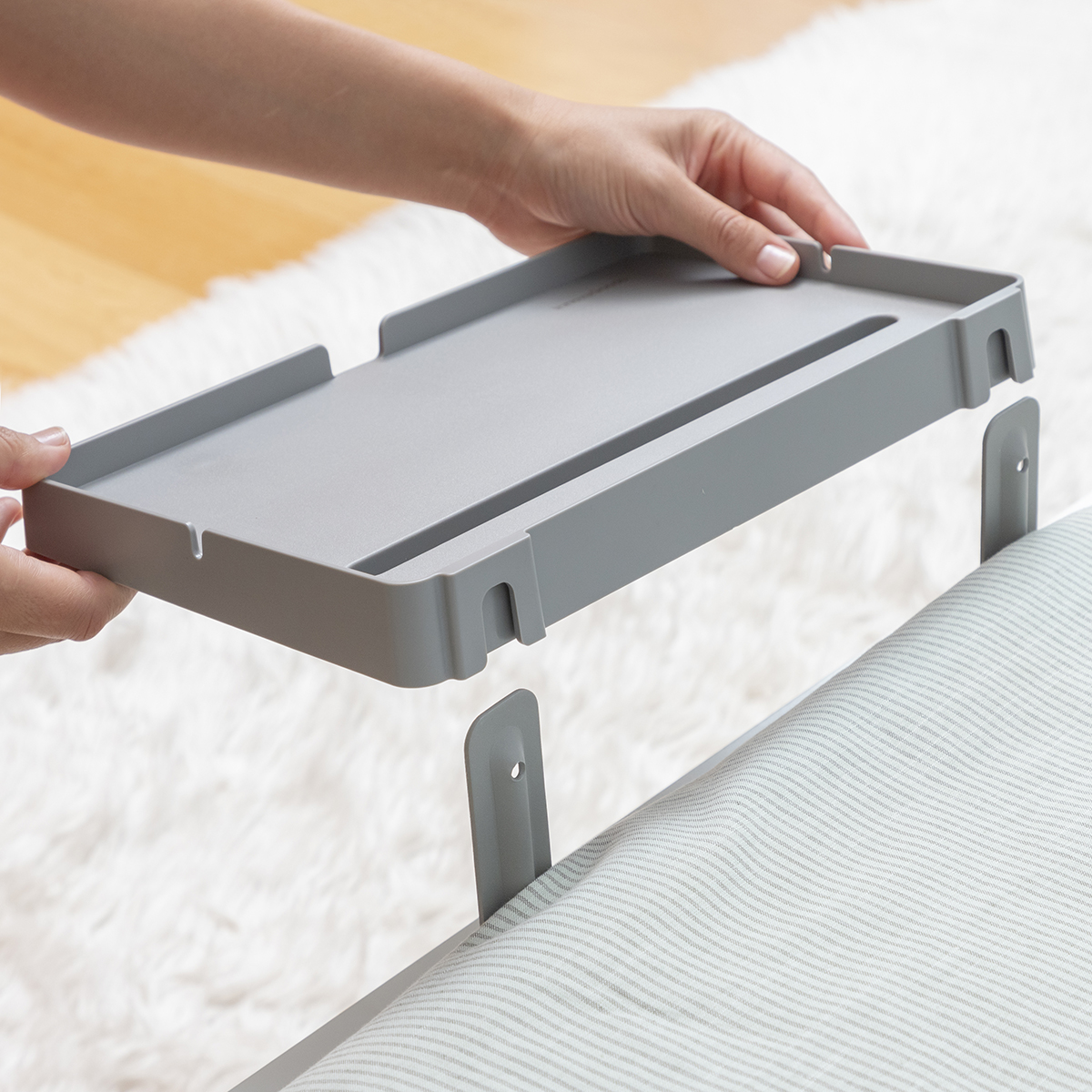 Mani che assemblano un vassoio grigio e staffe a L su un letto con lenzuola a righe, primo piano su un tappeto bianco.