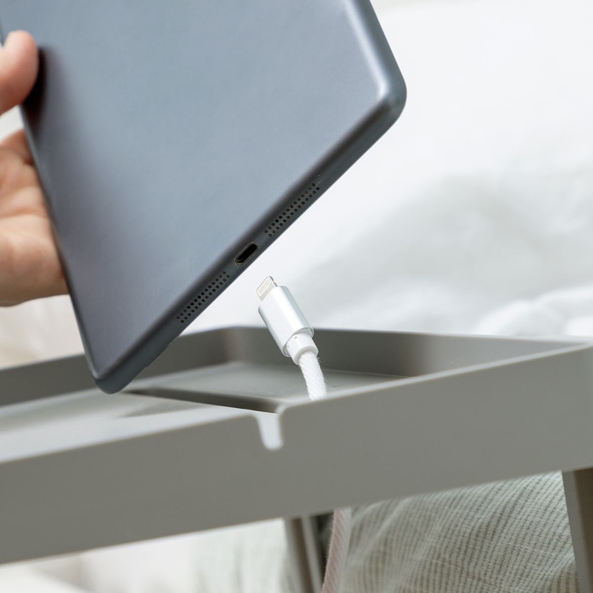 Una mano che collega un cavo di ricarica bianco a un tablet grigio su un vassoio grigio.