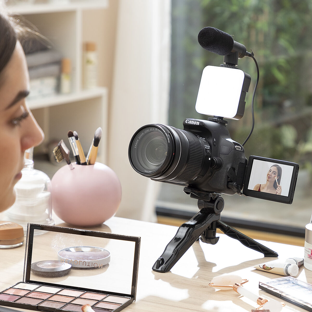 Mujer grabando con cámara, luz y micrófono. Maquillaje y espejo en la mesa.