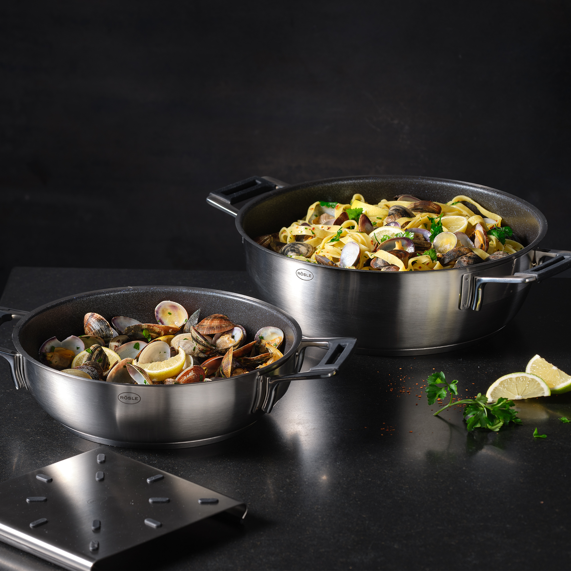 Zwei Pfannen mit Essen. Eine mit Pasta und Muscheln, die andere nur mit Muscheln, auf schwarzem Tisch.