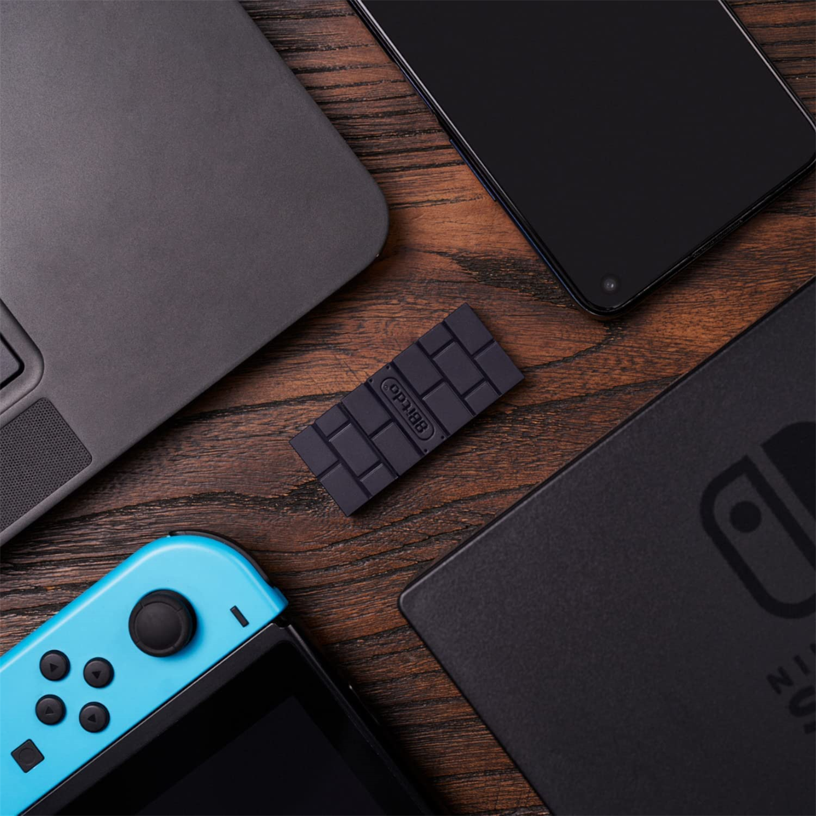Schwarzes, ziegelformiges Gerät, umgeben von Laptop, Telefon und Spielekonsole.