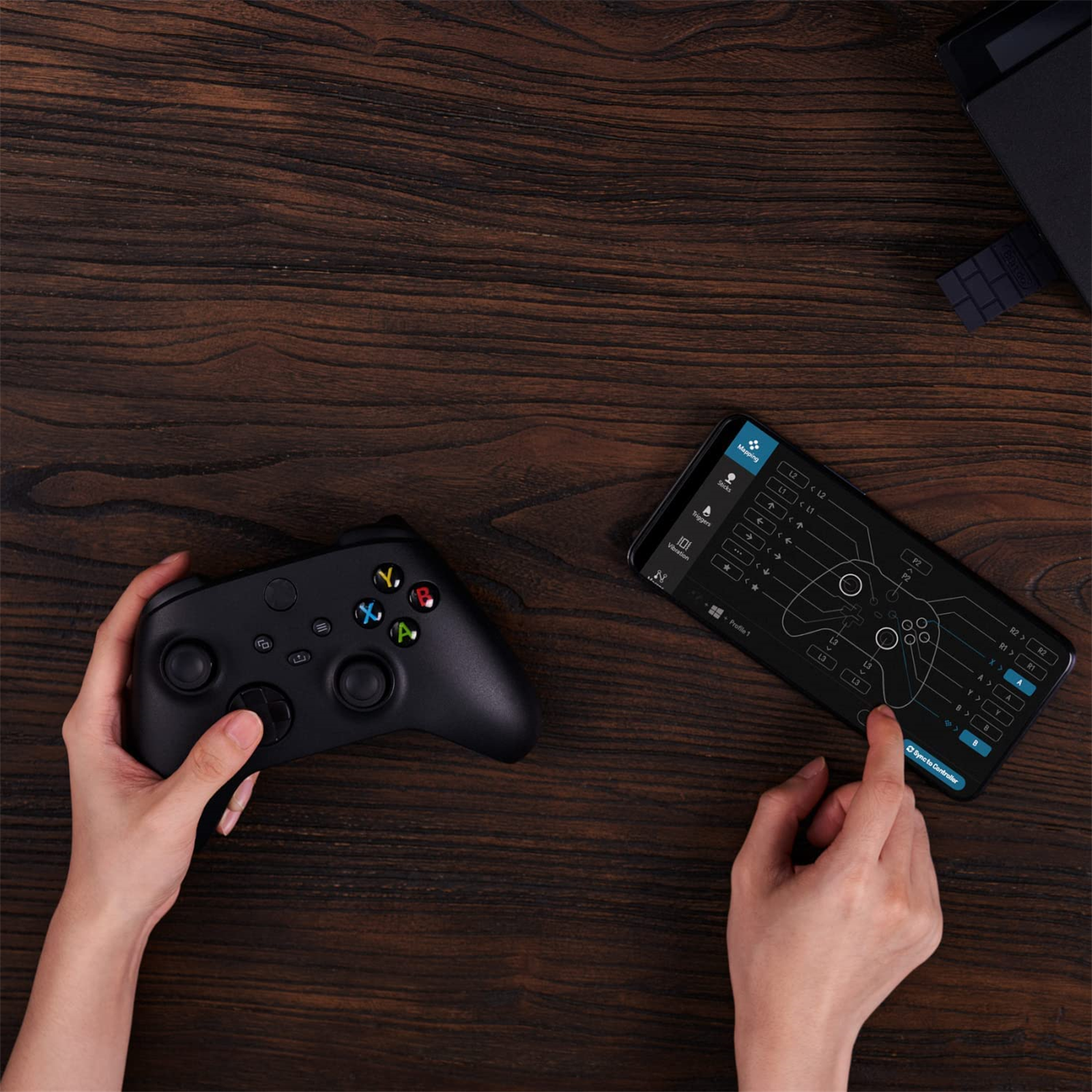 Hände halten einen schwarzen Gamecontroller mit einem Telefon daneben, das eine Spieloberfläche anzeigt.