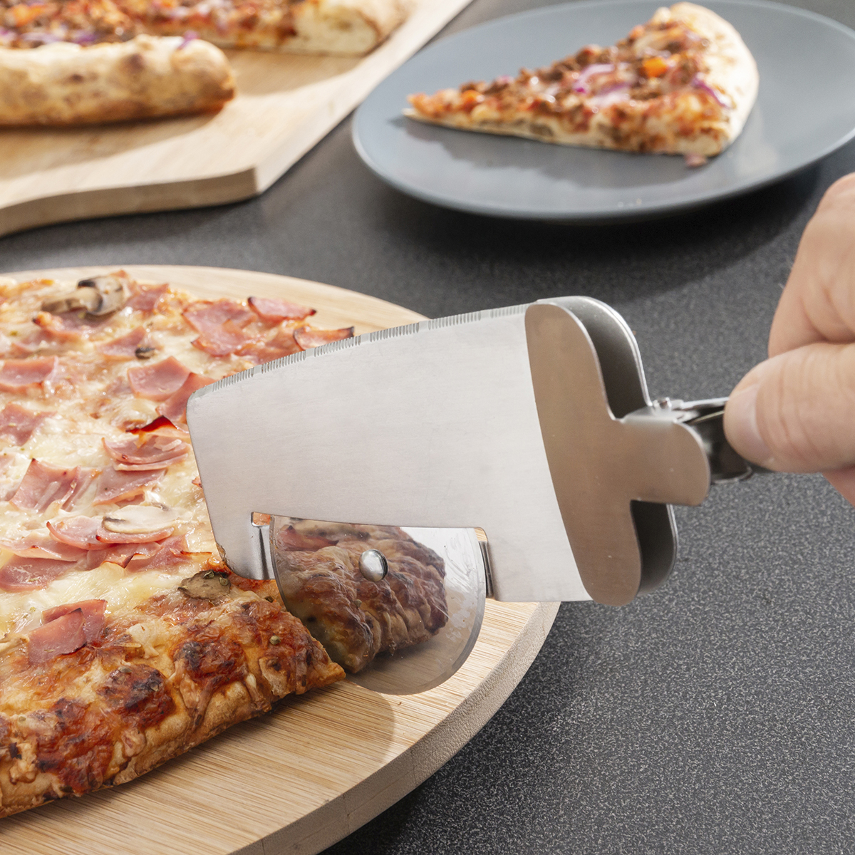 Una mano taglia la pizza con un taglia pizza su un tagliere di legno. Un'altra pizza su un piatto grigio è sullo sfondo.